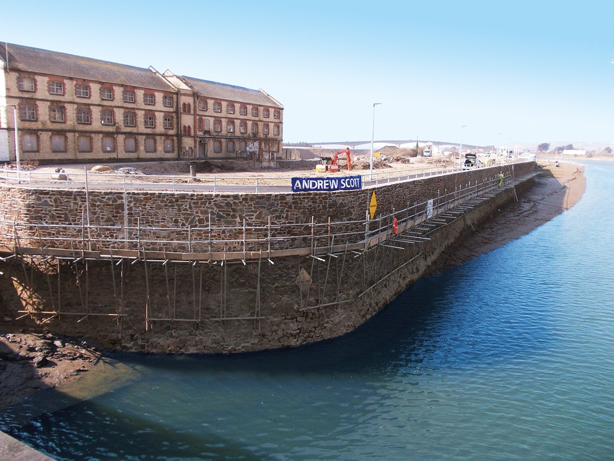 A construction site along a waterfront with scaffolding, a stone wall, and a sign that reads "Andrew Scott." There are old brick buildings behind, construction equipment, vehicles, and workers on site.