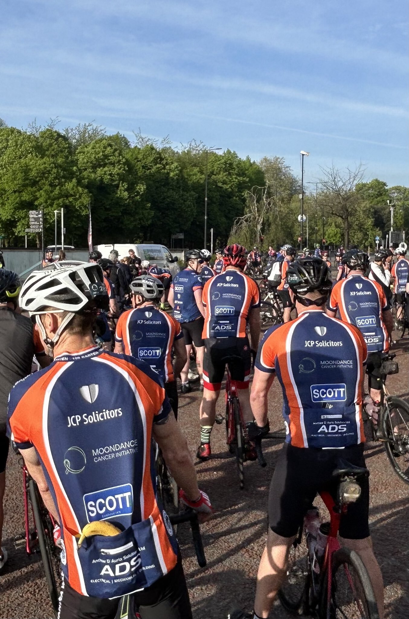 Group of cyclists gathered outdoors, wearing jerseys with various sponsor logos, preparing for a ride on a sunny day with blue skies and green trees in the background.