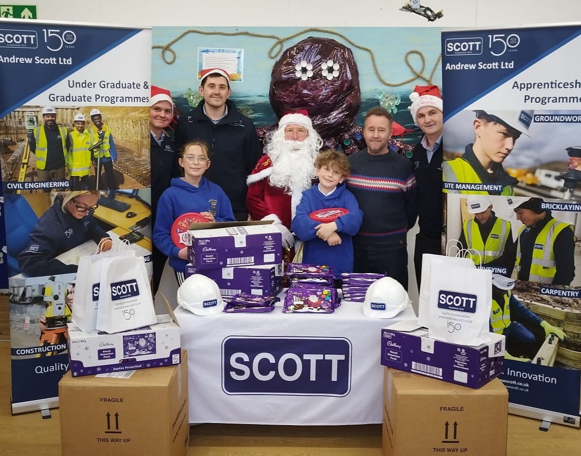 Group of people, including children and Santa Claus, standing behind a table with gifts and boxes at a company event, Christmas decorations and banners for Scott in the background.