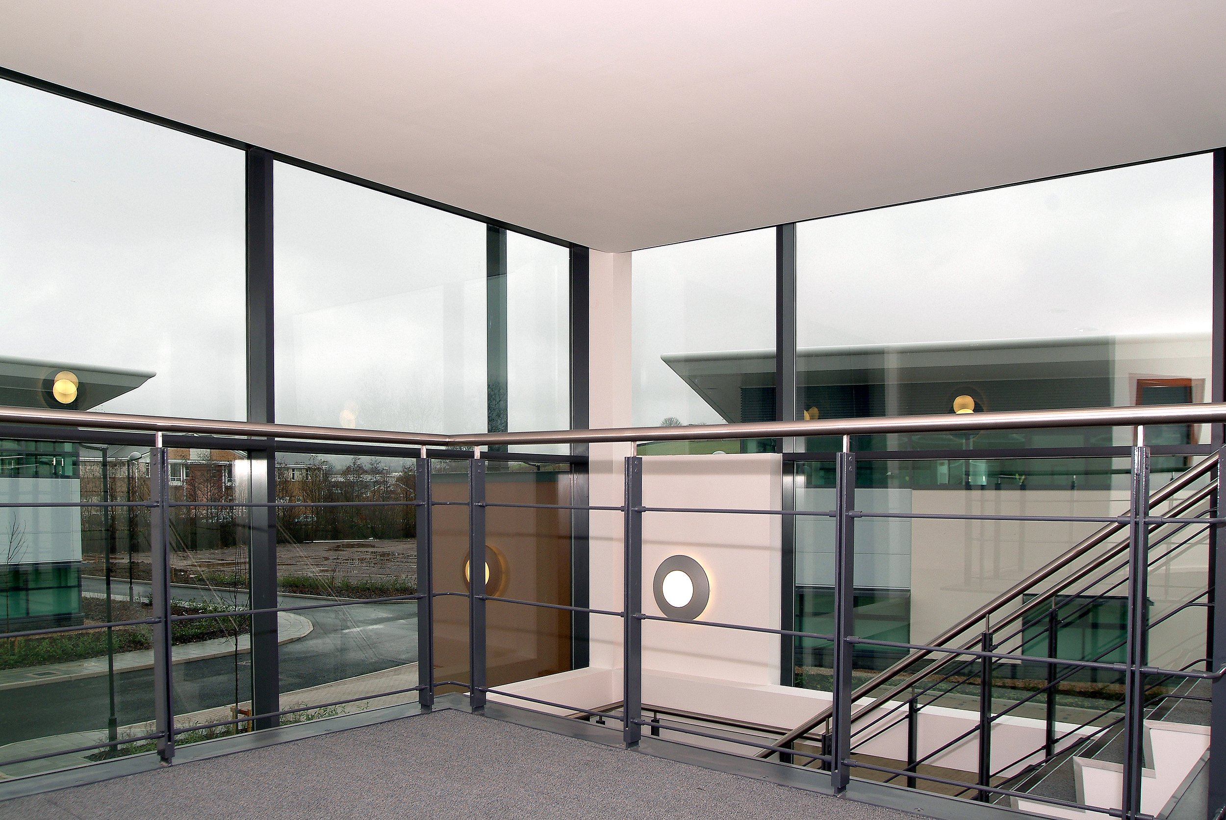 Interior view of a modern building with large glass windows, metal railings, and an outdoor scene visible through the windows, including a parking lot and additional architectural structures.