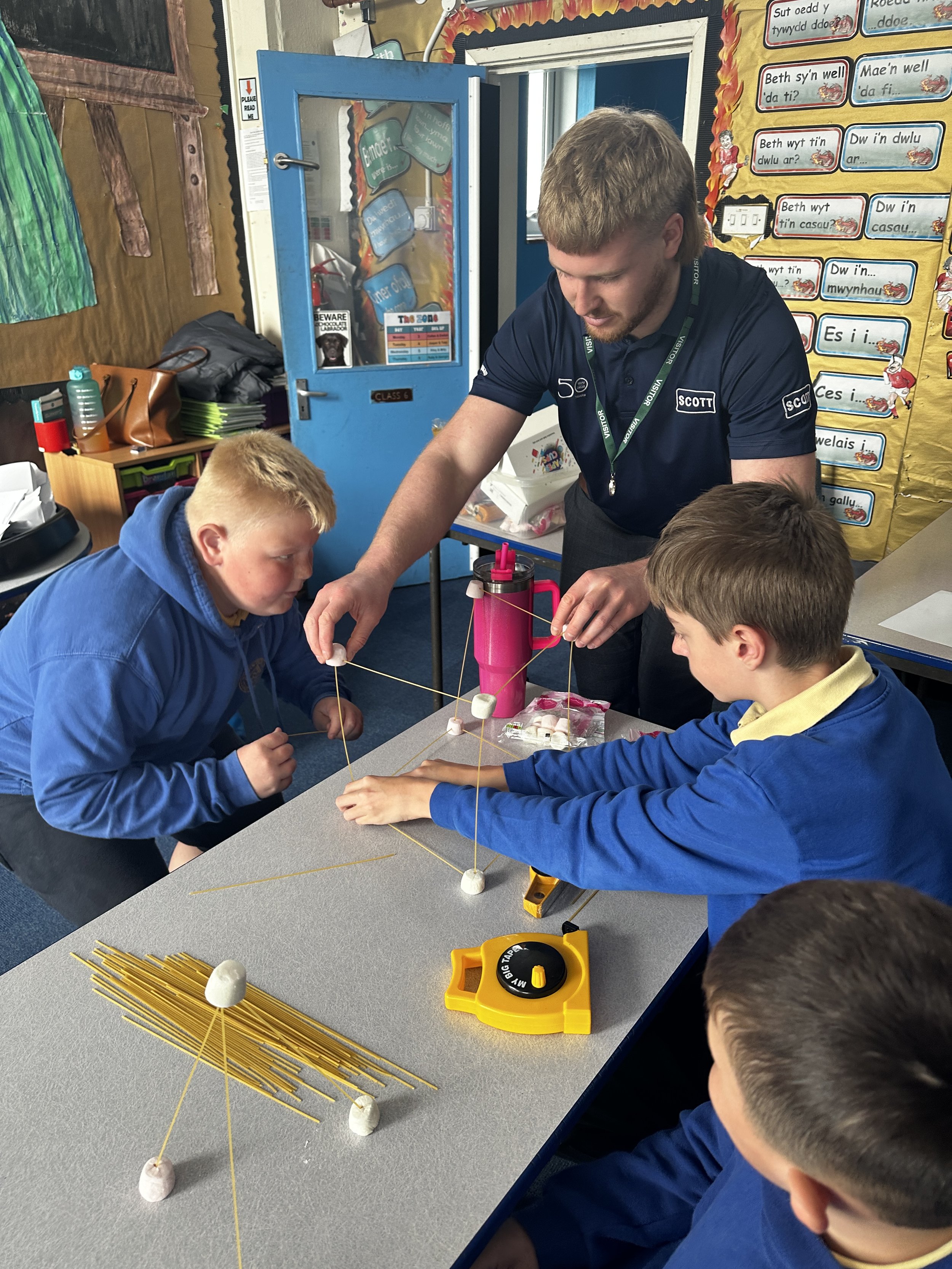 A classroom setting where a teacher is supervising three students as they work on a project involving marshmallows and uncooked spaghetti sticks to create structures. The teacher and students are focused on constructing a geometric shape. There are s