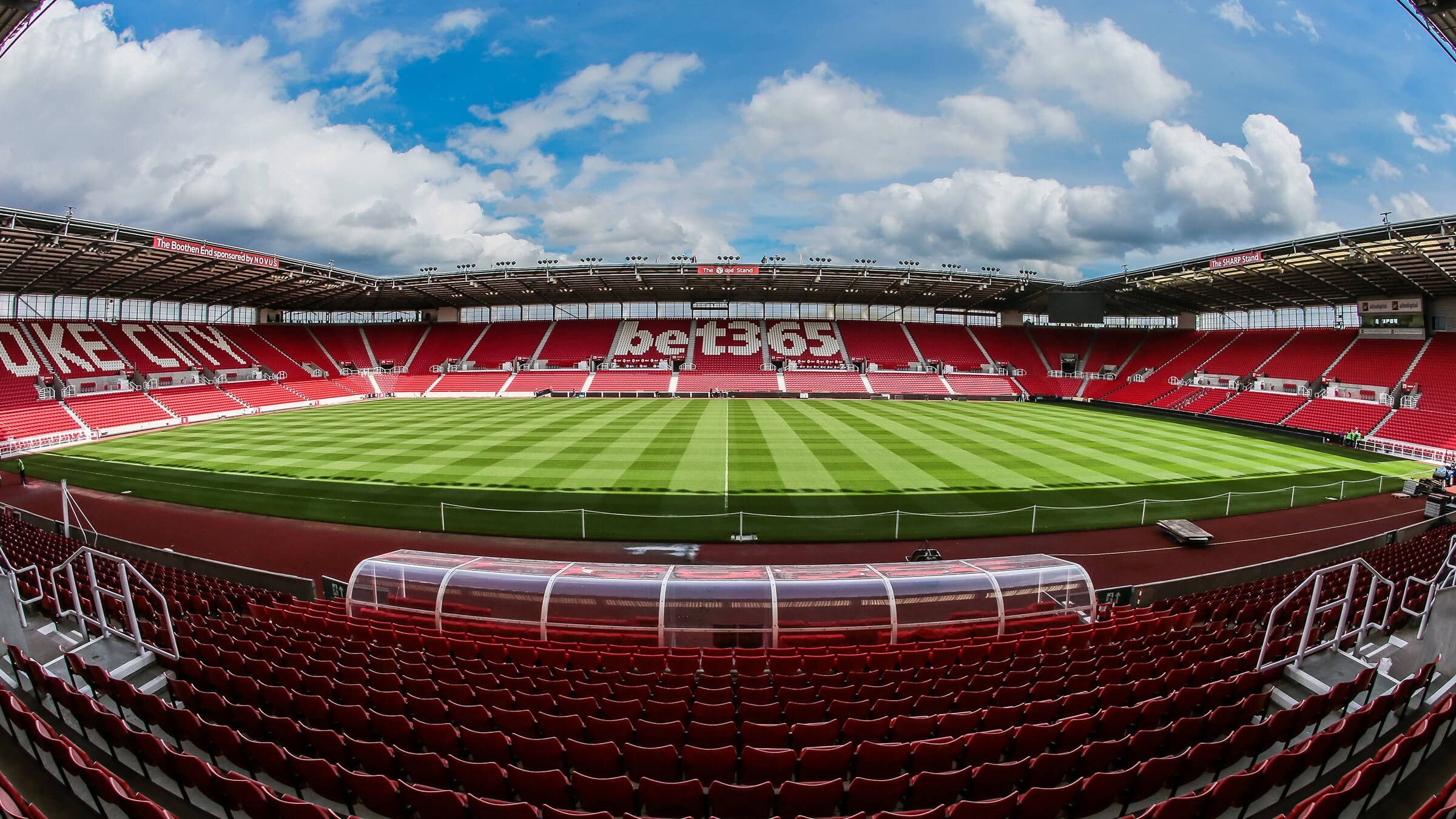 Empty soccer stadium with green field, red seats, and a partially cloudy sky.