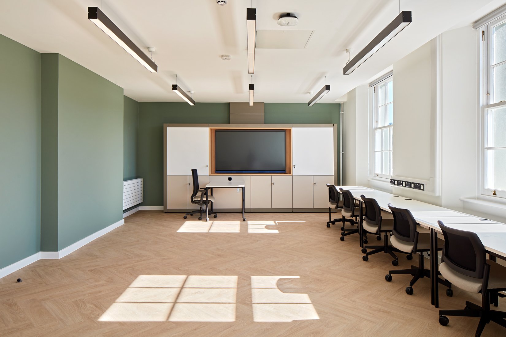 Modern classroom with green walls, large windows, desk with chairs, and interactive whiteboard.