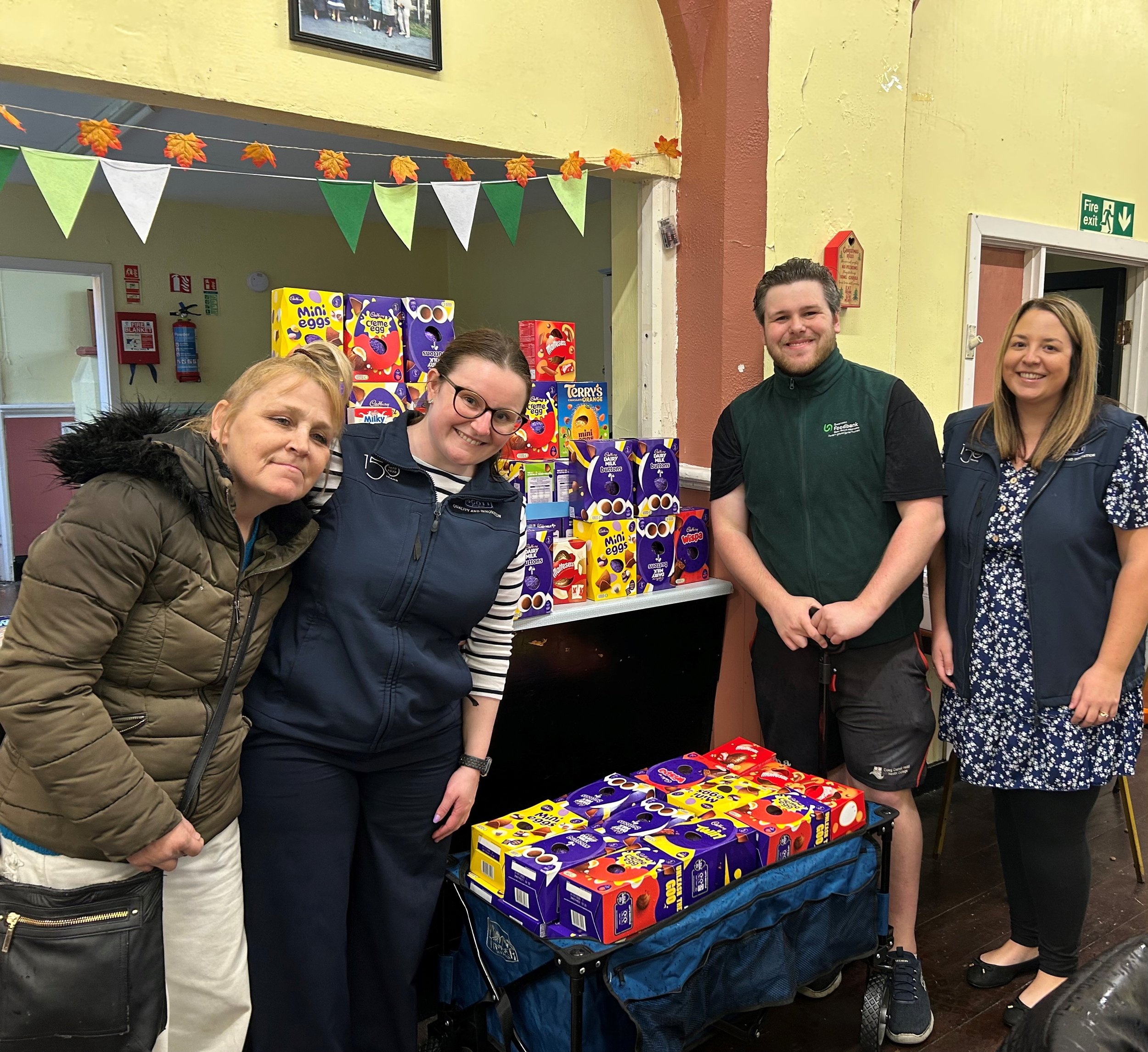 Four people standing behind a table and shelves filled with Easter chocolate, smiling for the camera in a decorated indoor space.