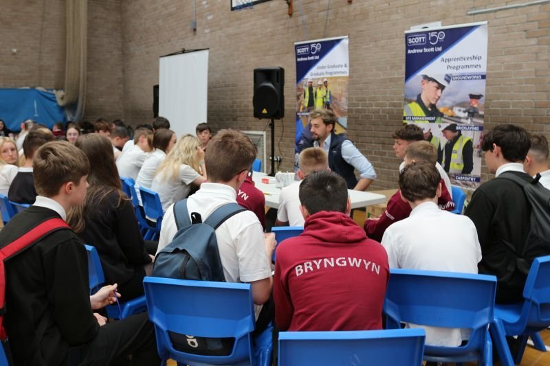 Students listening to a speaker at a college or career fair in a large room with posters about apprenticeship programs and skills like management, broking, carpentry, and plumbing.