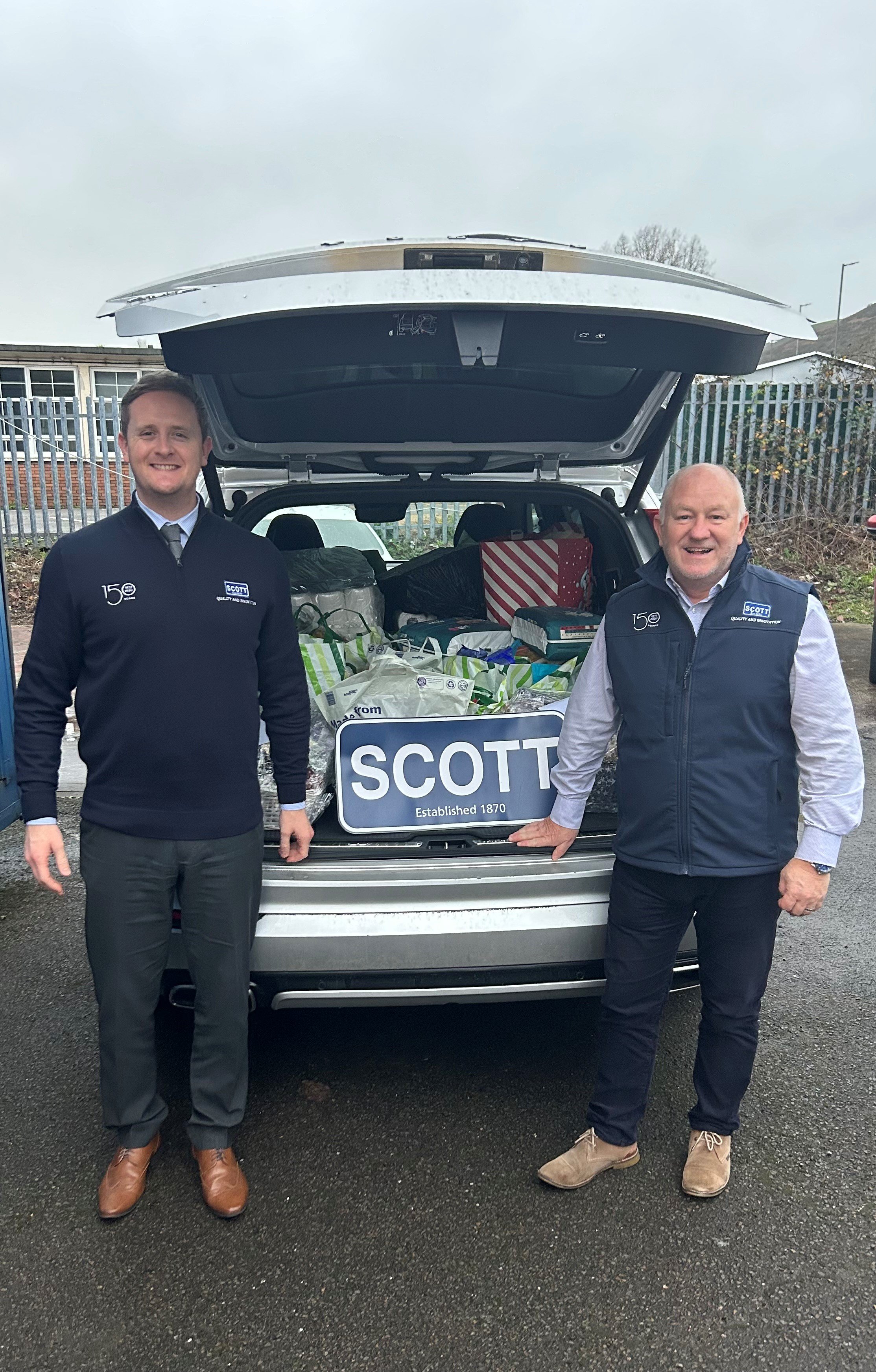 Two men standing next to an open car trunk filled with gifts and a Scott sign, outdoors on a cloudy day.