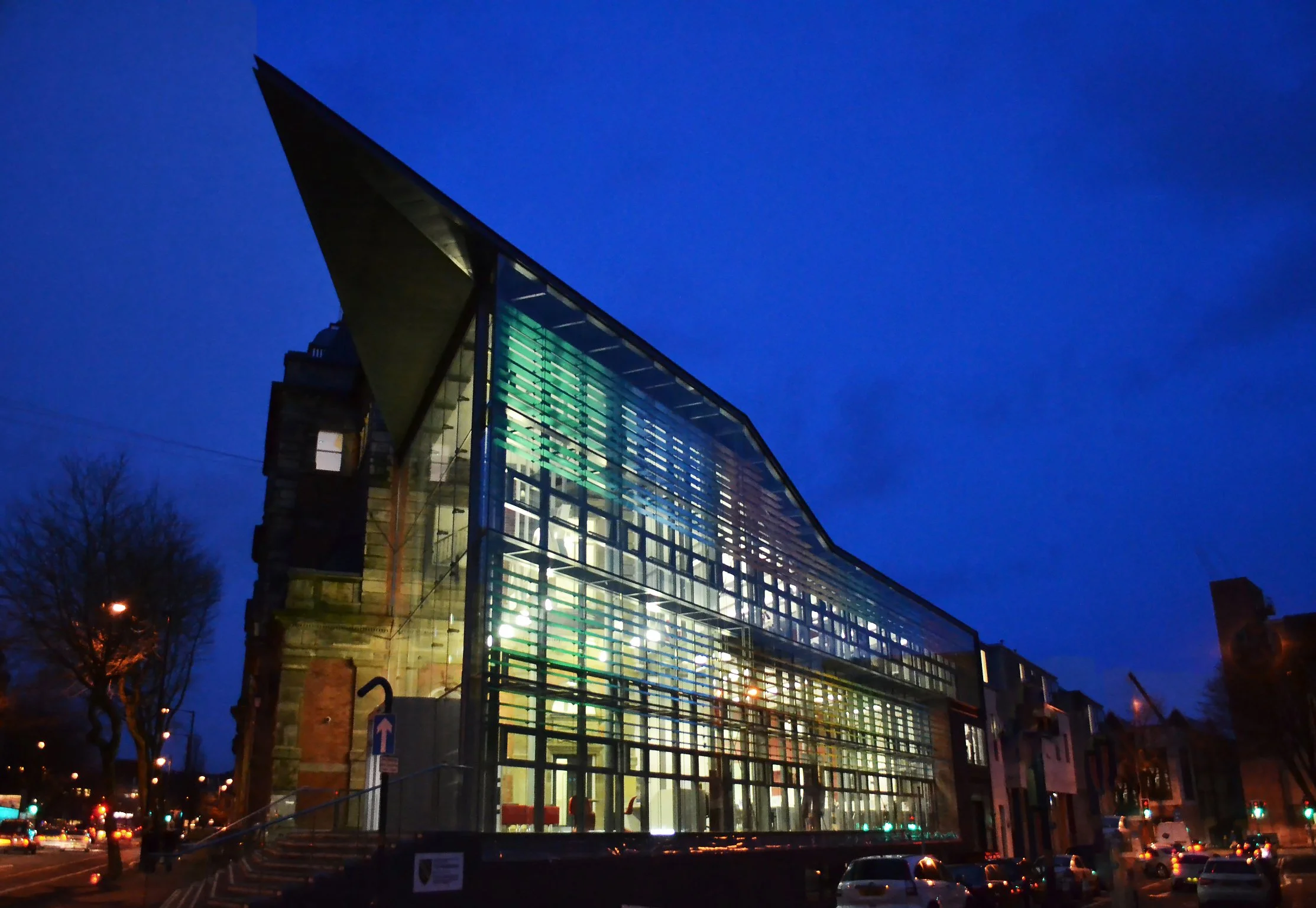 A modern glass building illuminated at night, with city lights and parked cars visible in the foreground.