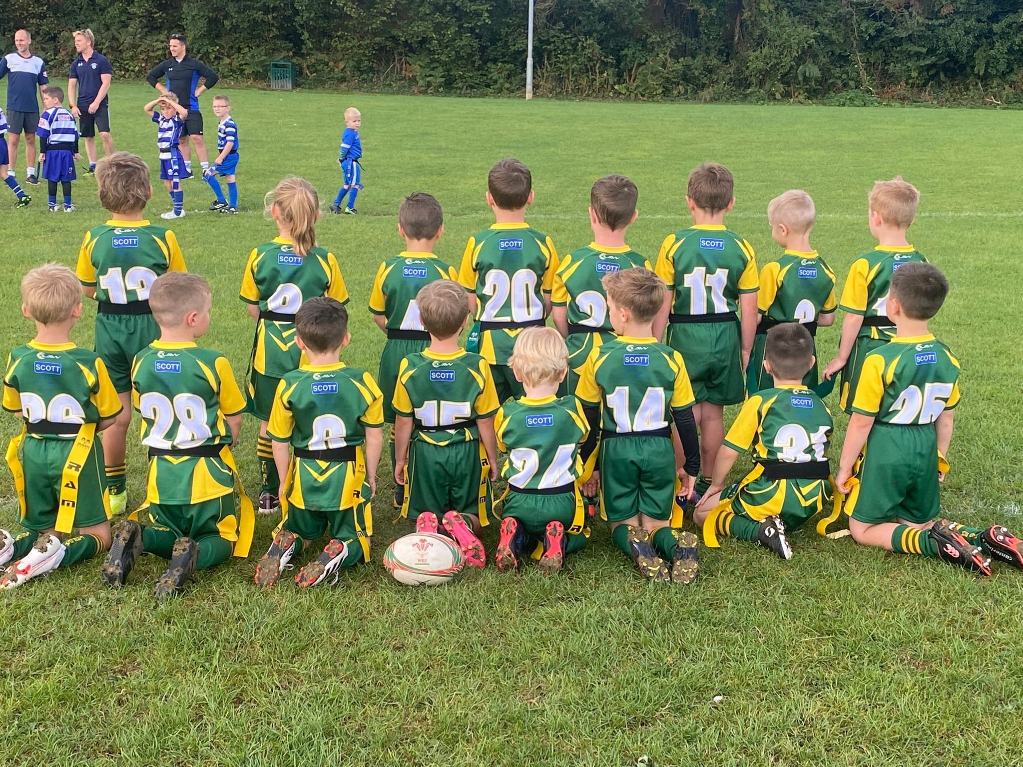 Group of young children in green and yellow rugby uniforms kneeling on grass, with some players standing in the background, on a rugby field.