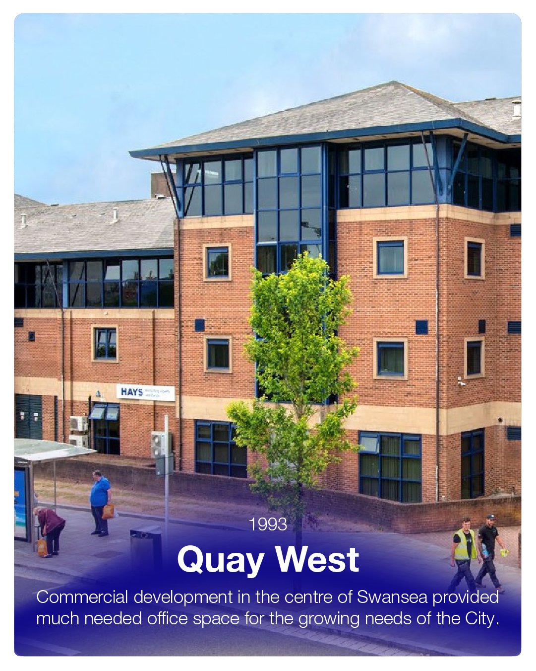 Photograph of a modern, multi-story office building with brick and glass exterior, and a tree in front, located at Quay West in Swansea.