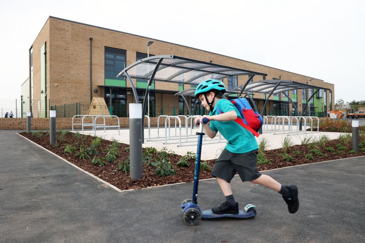 Child riding a scooter in front of a modern school building.