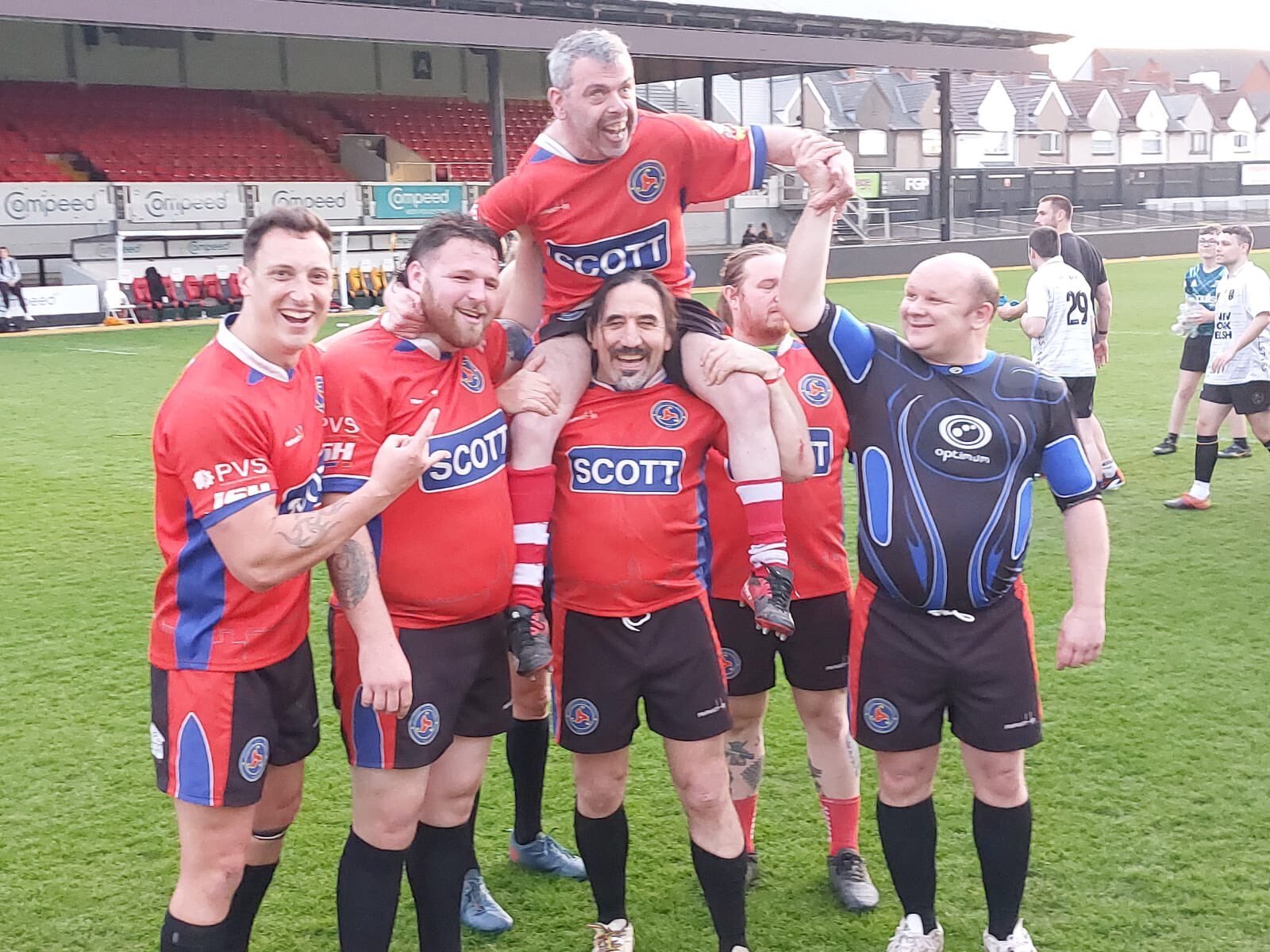 A group of rugby players celebrating on the field, with one player being lifted on others' shoulders, all smiling and pointing, wearing red and blue jerseys.