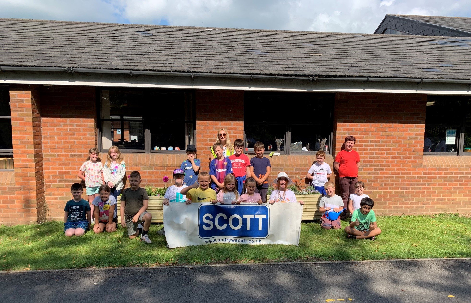 Group of children and two adults posing outside a brick building with a banner that reads 'SCOTT' and a website URL, on a sunny day.