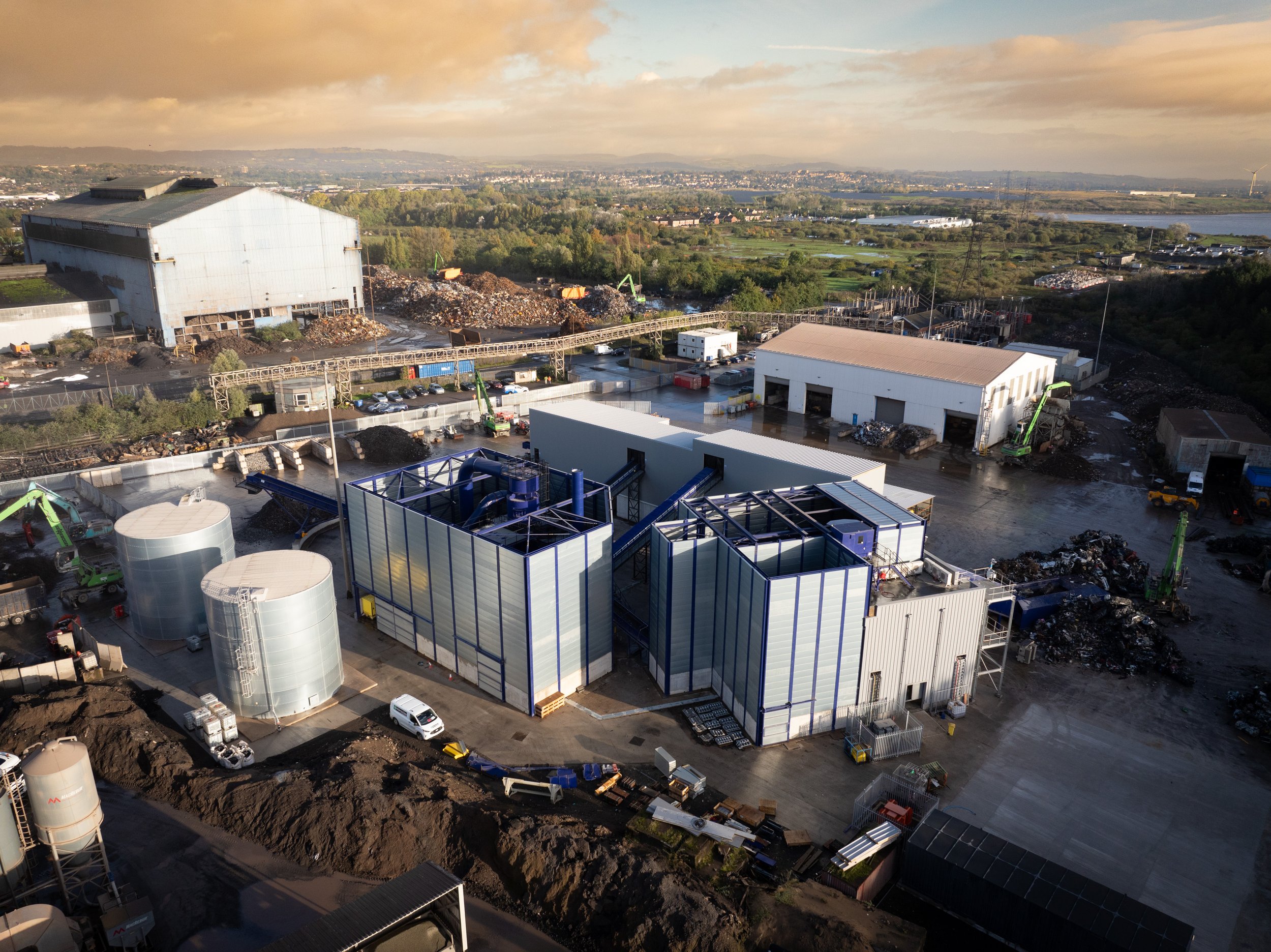 Aerial view of industrial plant with warehouses, storage tanks, and machinery on a large site, surrounded by piles of materials and greenery in the background.