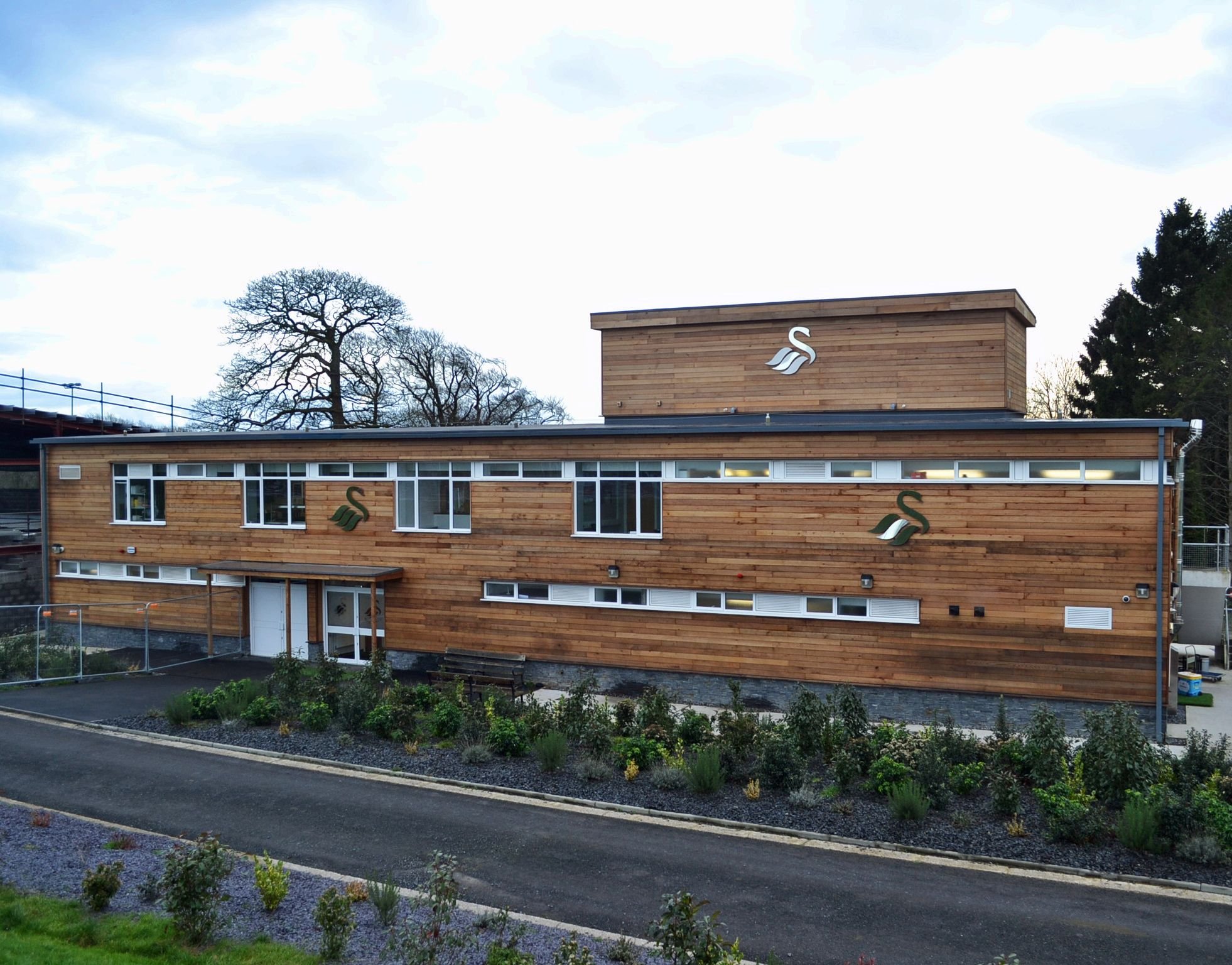 Modern wooden building with swan logos on the facade, surrounded by landscaped gardens and trees, cloudy sky in background.
