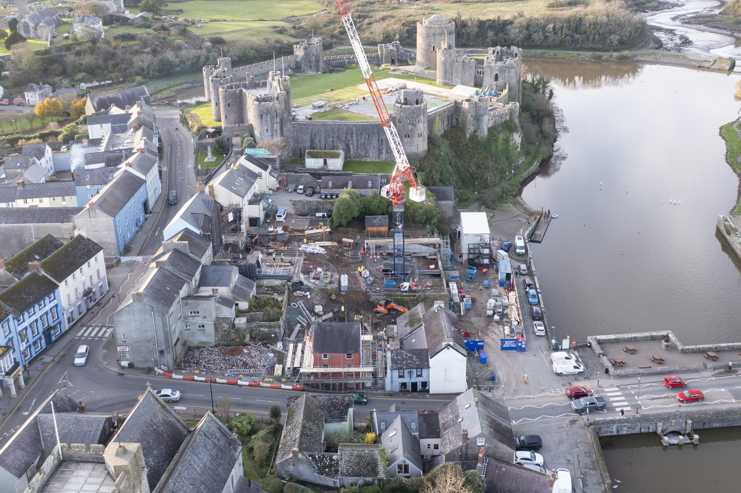 Aerial view of a construction site near a historic castle and river. Surrounding buildings, roads, and greenery are visible.
