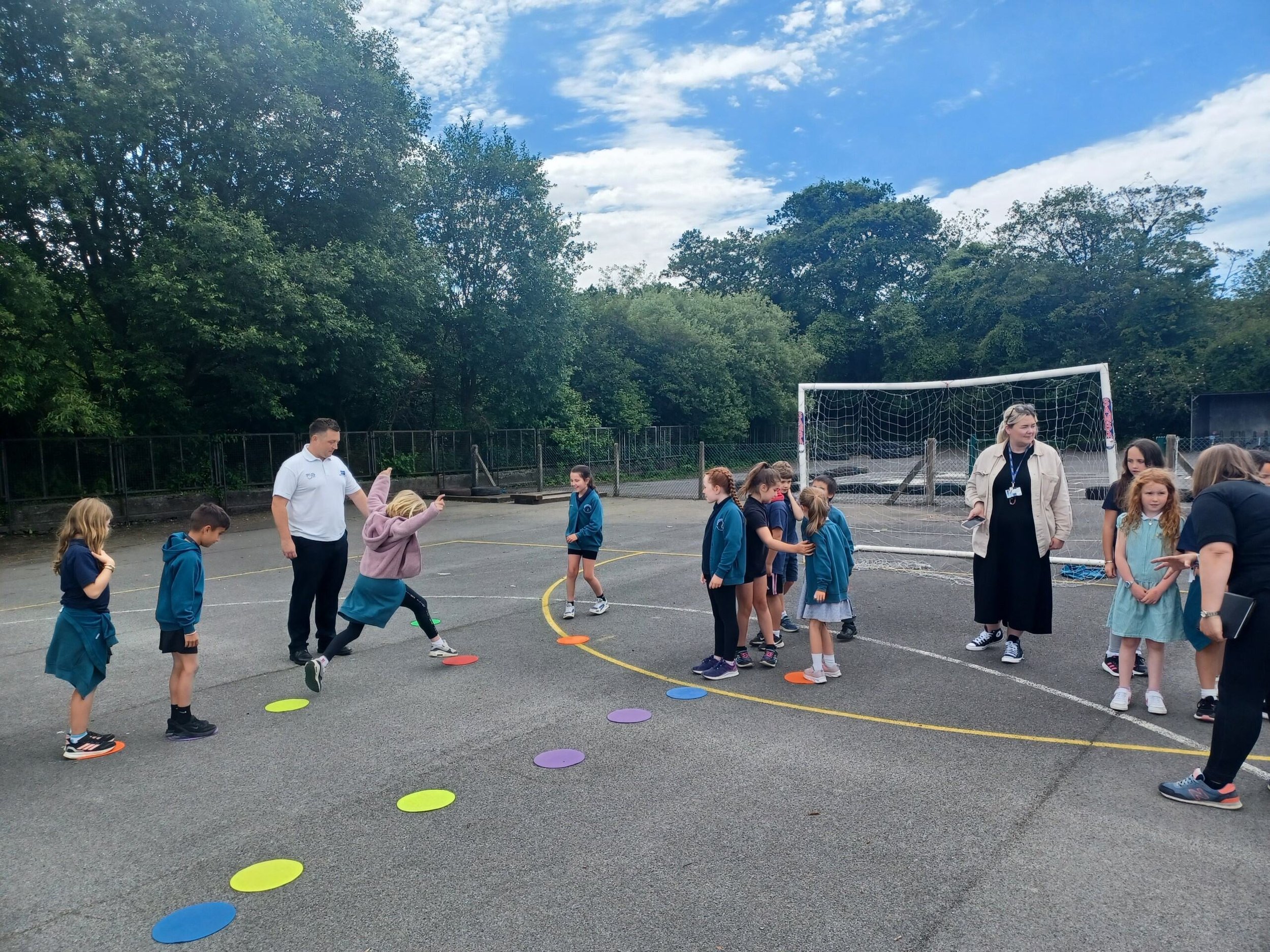 Children participating in outdoor activities on a school playground with teachers supervising, colorful cones on the ground, and a soccer goal in the background.