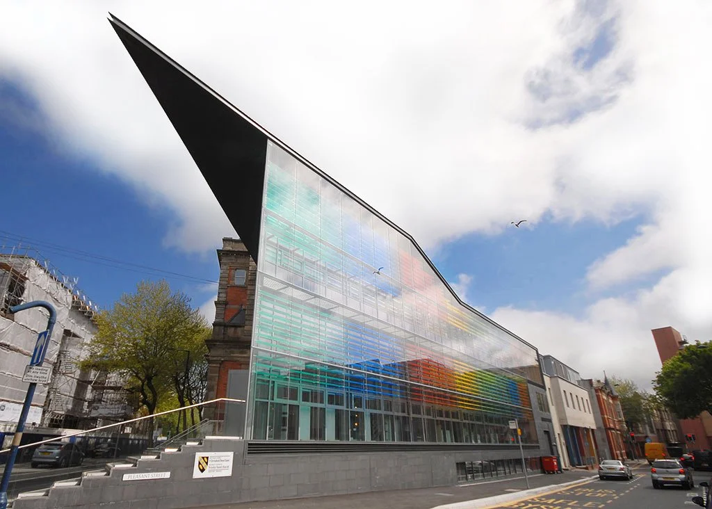 Modern building with a glass facade and colorful rainbow stripes, situated in an urban area with trees and parked cars.