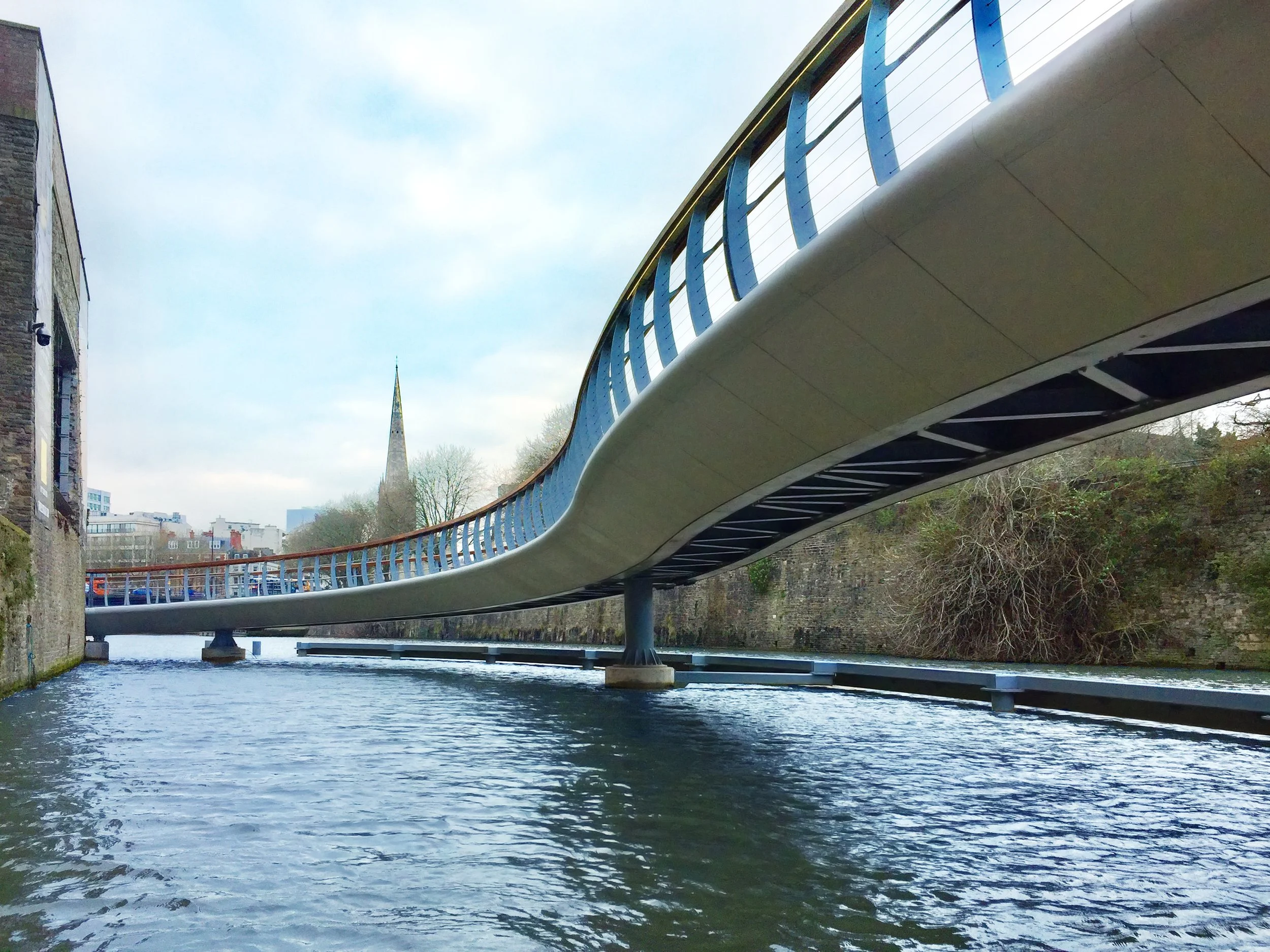 Modern pedestrian bridge spanning over a river with a cityscape and church steeple in the background.