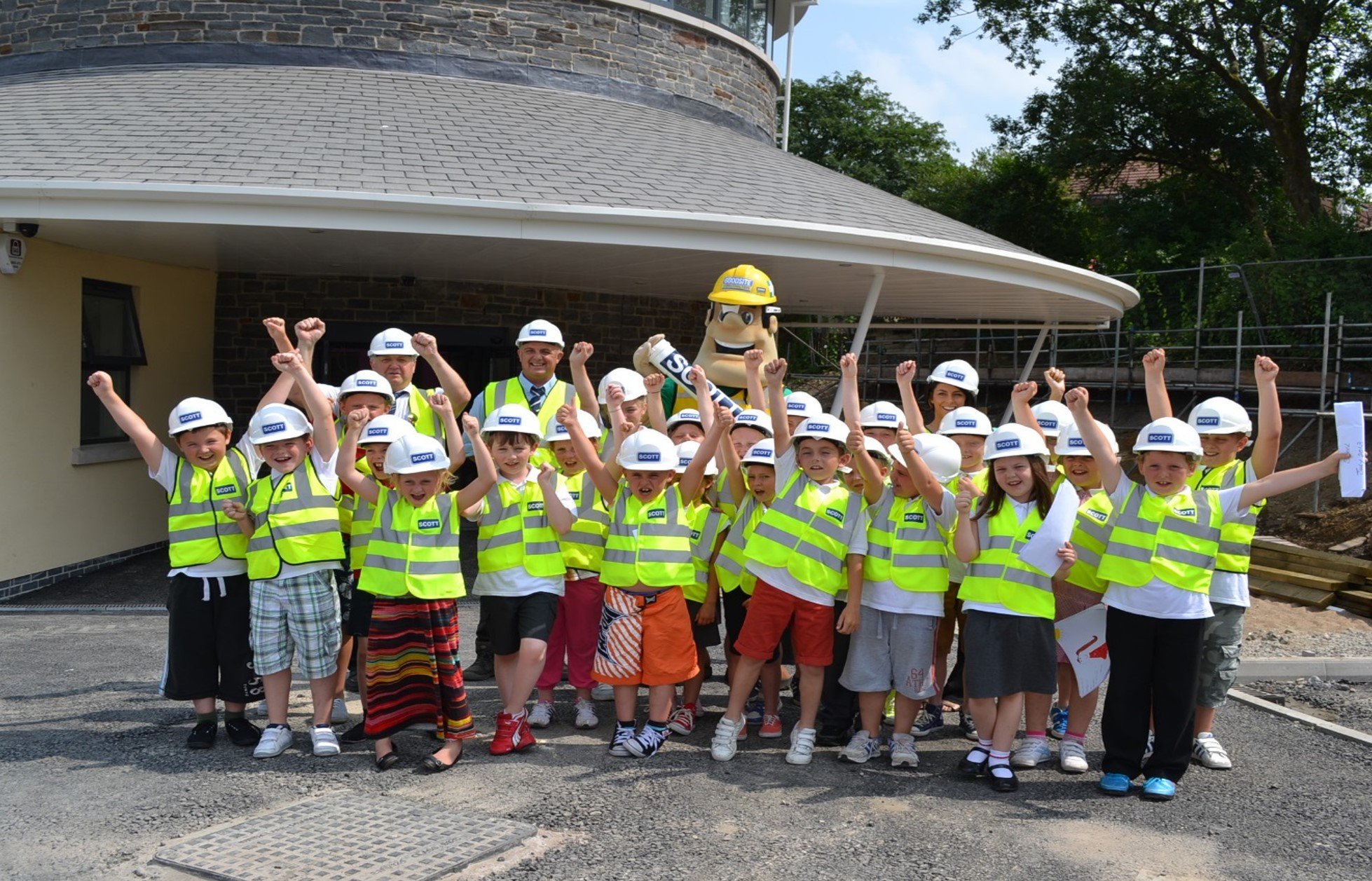 Group of children wearing yellow safety vests and construction helmets, celebrating outdoors with a mascot dressed as a construction worker.