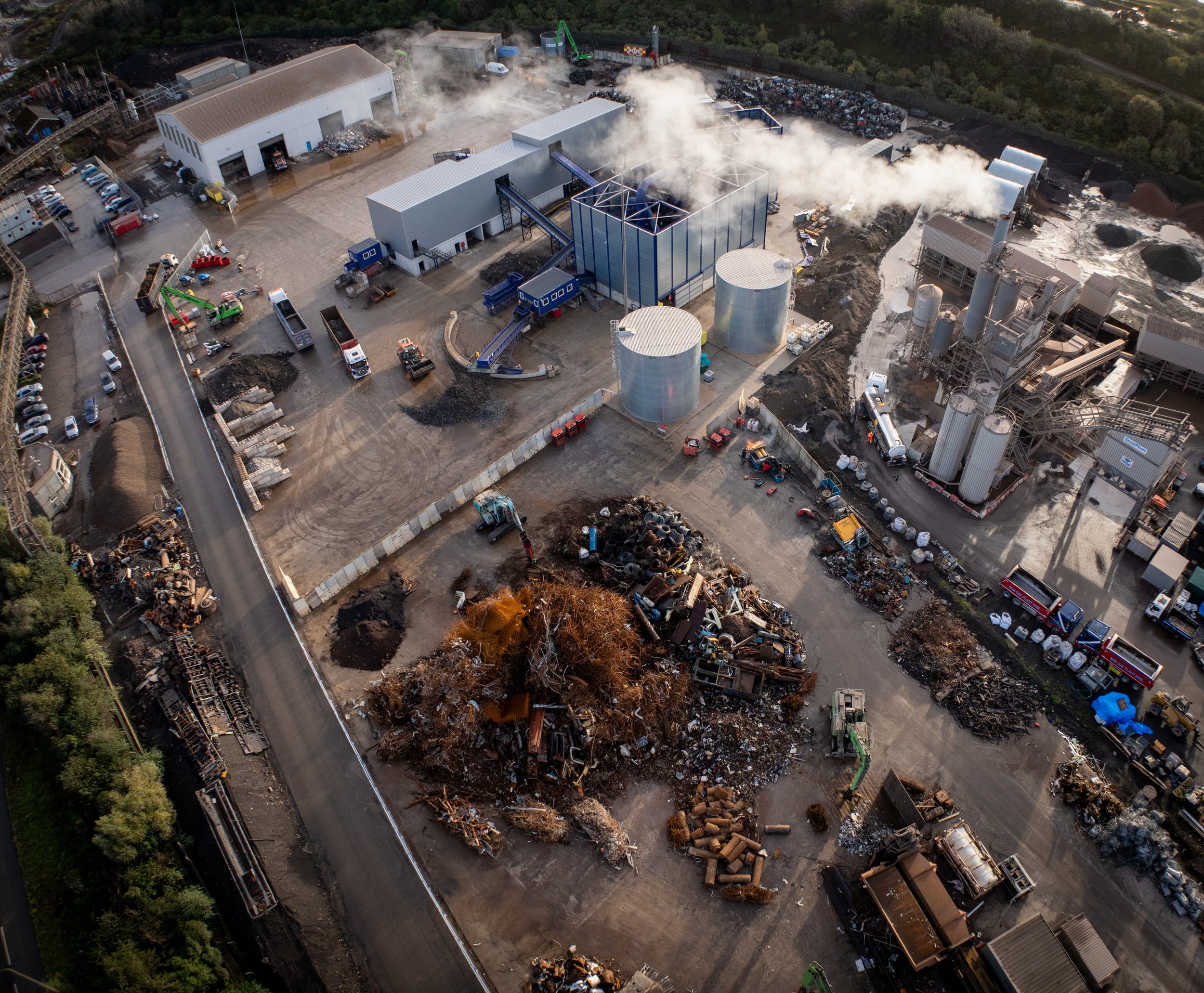 Aerial view of an industrial recycling facility with scrap metal piles, warehouses, trucks, and machinery. Smoke is visible from processing equipment, and various materials are arranged around the site.