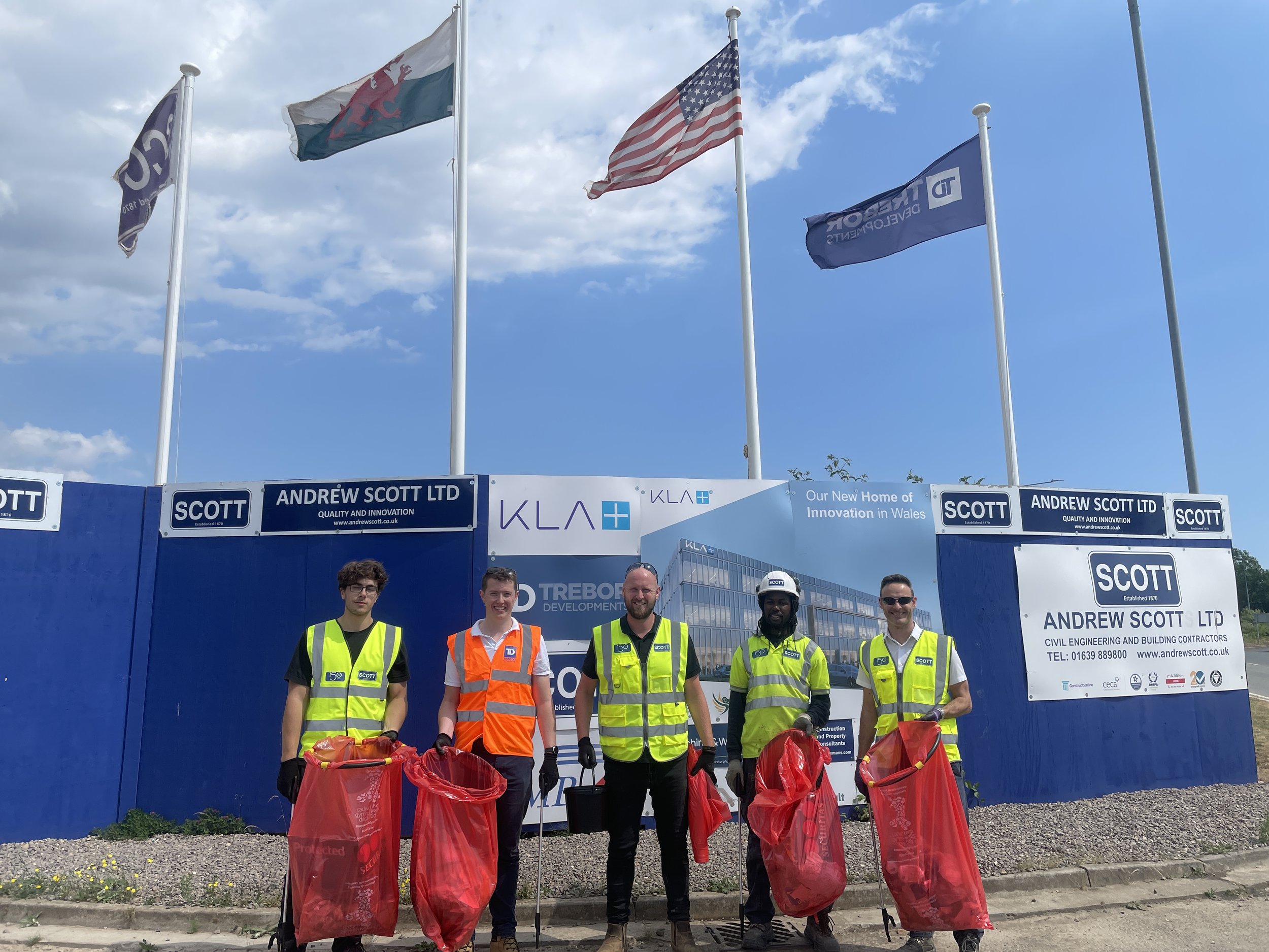 Five construction workers standing with trash bags in front of a construction site fence, with flags flying above. The workers are wearing safety vests and helmets.