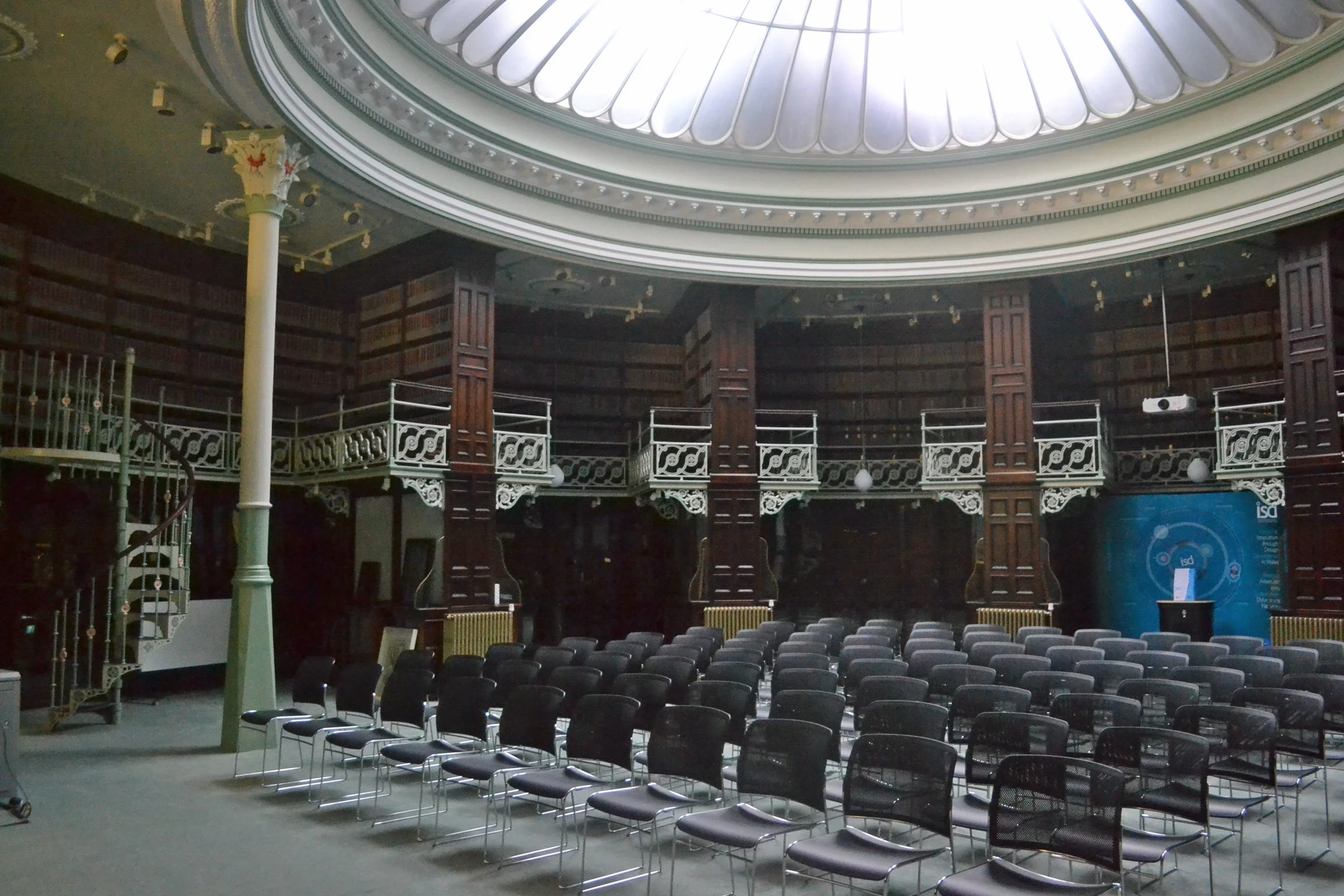 An auditorium with rows of black chairs, ornate white balcony railings, wooden columns, and a domed ceiling with skylight.