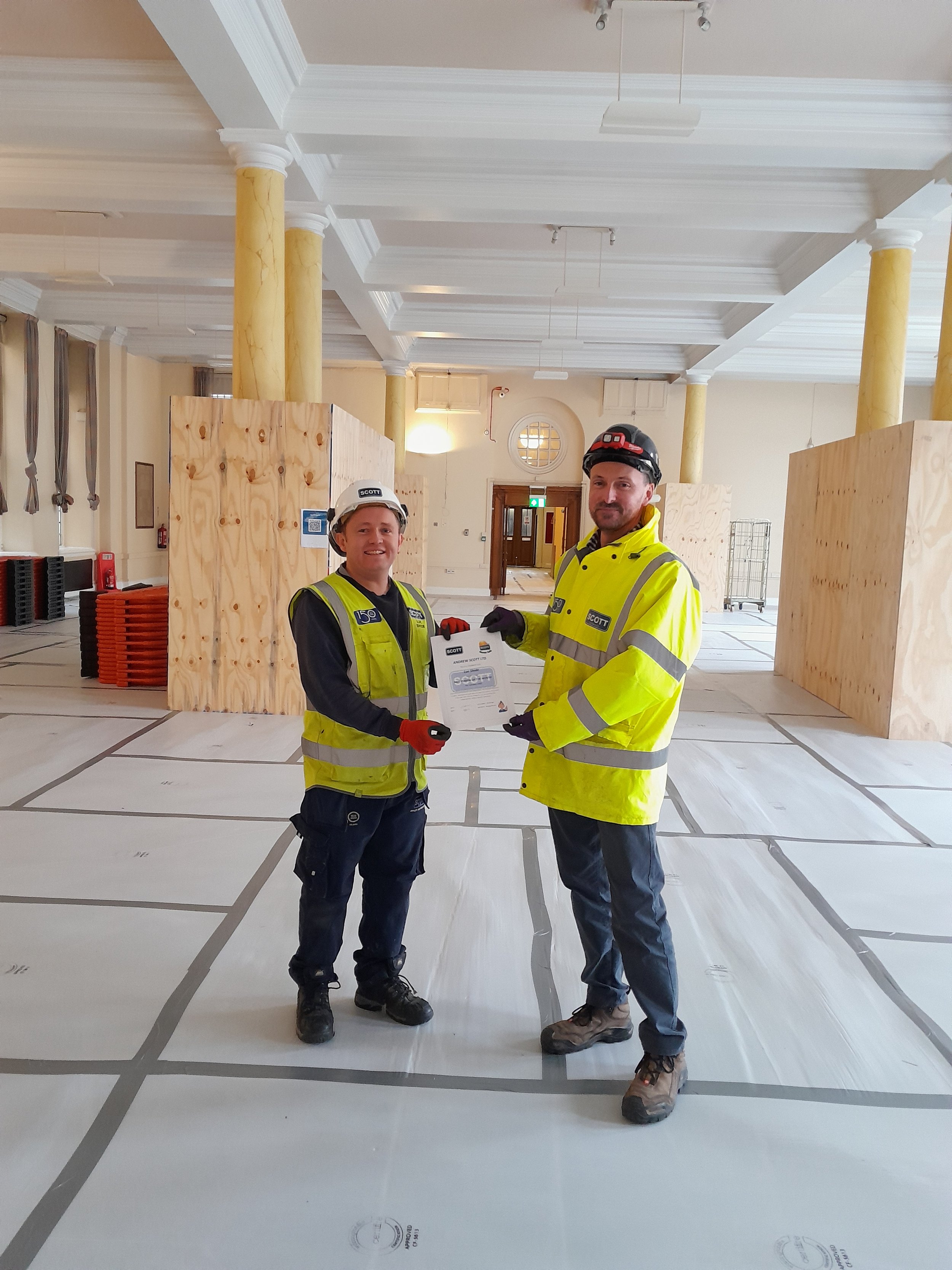 Two construction workers in high-visibility safety jackets and helmets standing inside a large room with scaffolding and construction materials, holding a document.