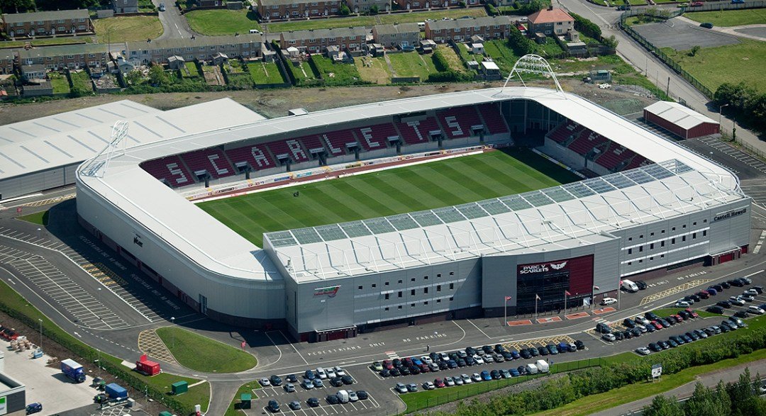 Aerial view of an outdoor sports stadium with a green field, covered seating, and "SCARLETS" spelled in the stands.