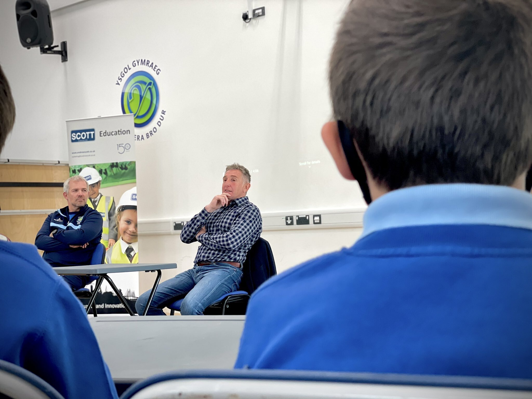 Two men speaking at a presentation or classroom in front of students. One man is sitting with his hand on his chin, wearing a checkered shirt and jeans. The other man is sitting with arms crossed, wearing a blue sports jacket. Students are wearing bl