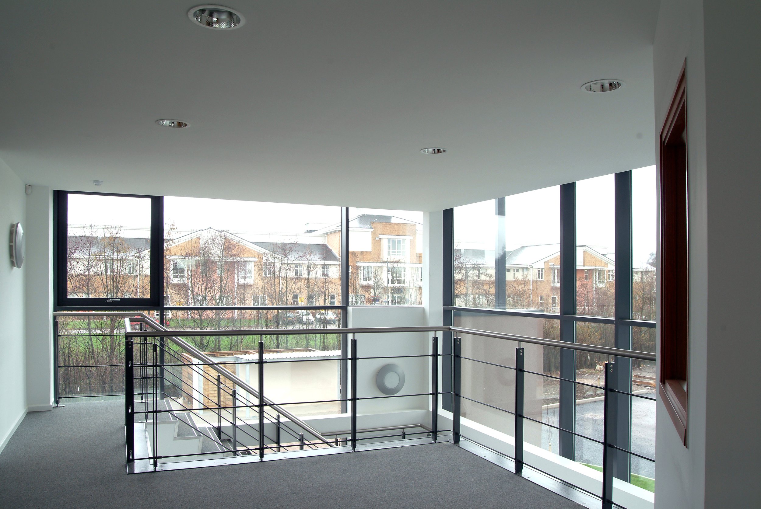 Interior view of a modern, well-lit upper-floor corridor with large windows overlooking a residential neighborhood with trees and houses, and a metal railing around an open staircase.