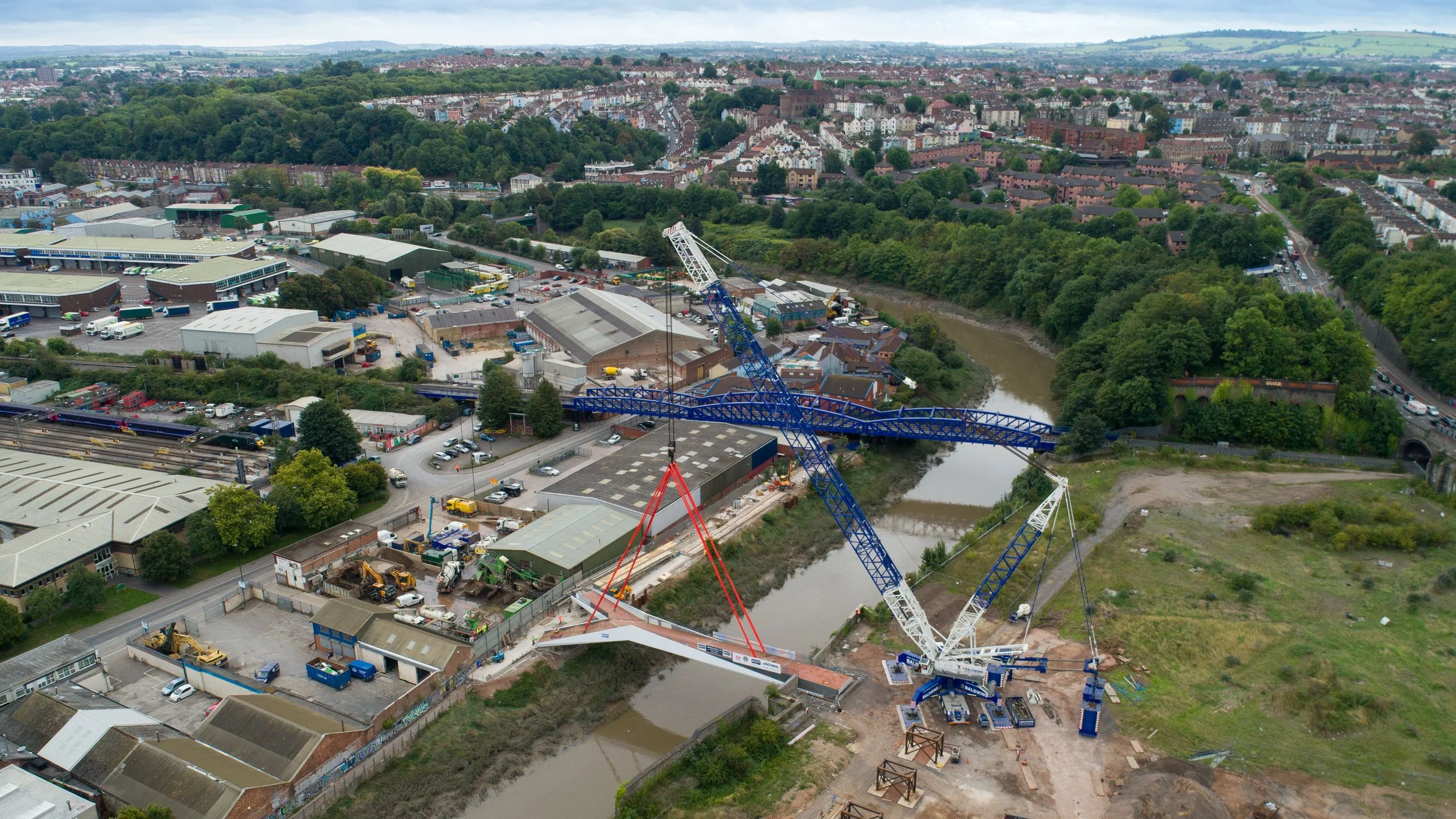 An aerial view of a construction site with a large blue and white crane installing a bridge span over a river, surrounded by an industrial area and a residential neighborhood.