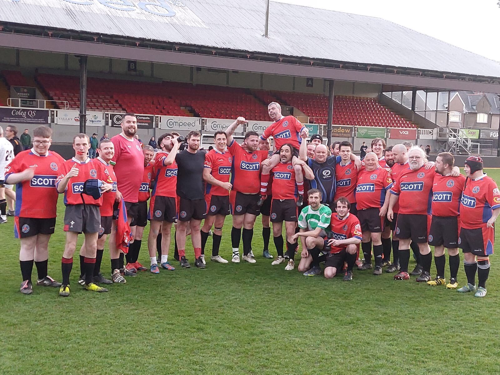 A group of rugby players and supporters celebrating on a field after a game, some wearing red jerseys with a blue logo and sponsor 'SCOTT', others in casual attire, in a stadium with empty red seats and advertising boards in the background.