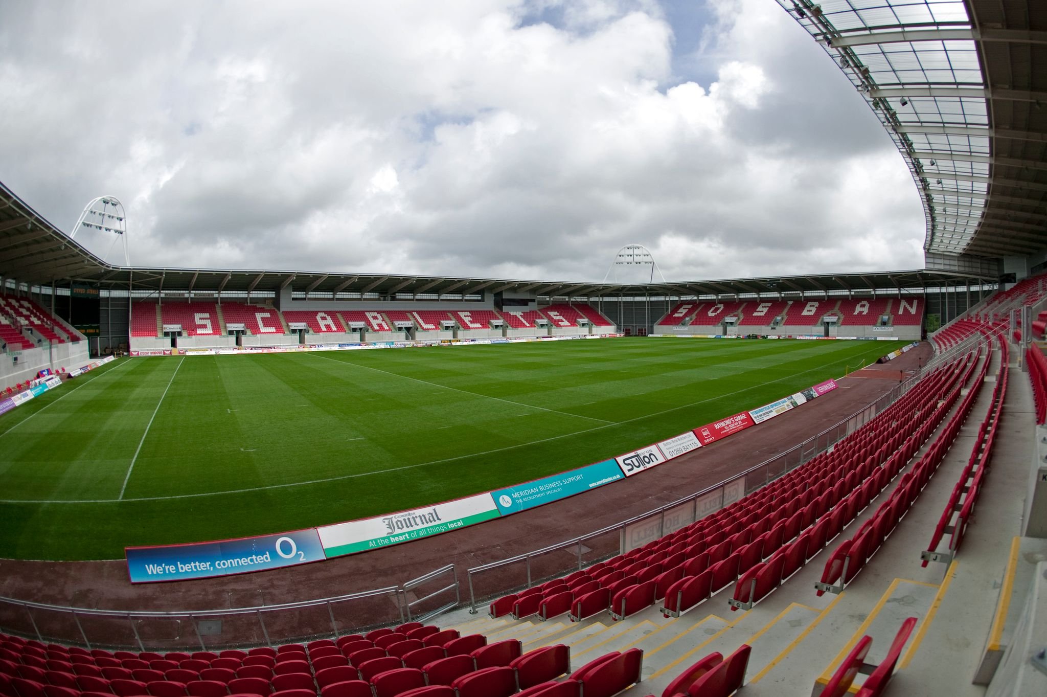 Empty rugby stadium with green field, red seats, and "Scarlets" lettering on stands under cloudy sky.