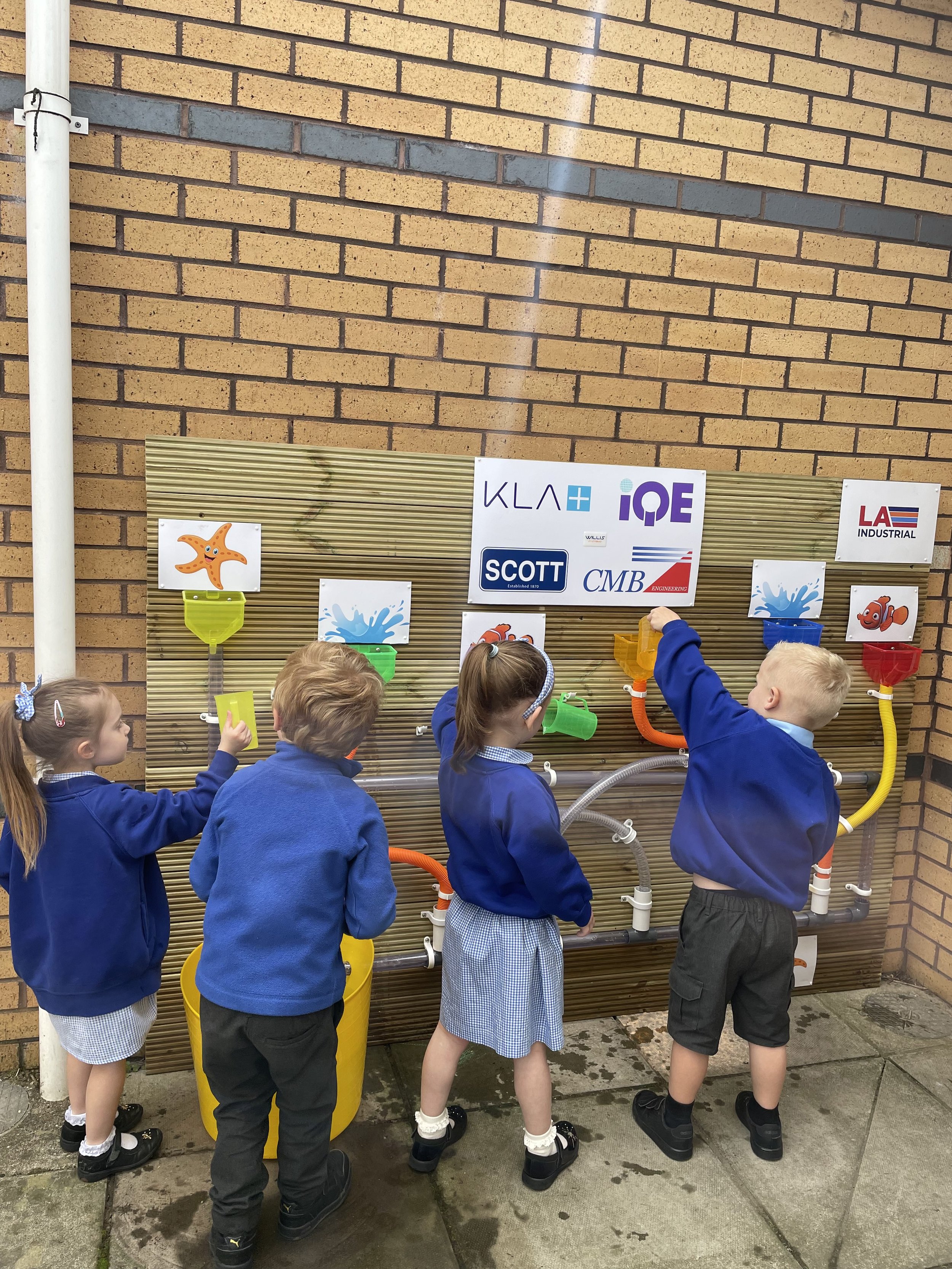 Four children in school uniforms engaging with a hands-on water activity display on a brick wall with company logos, including a starfish and fish illustrations.