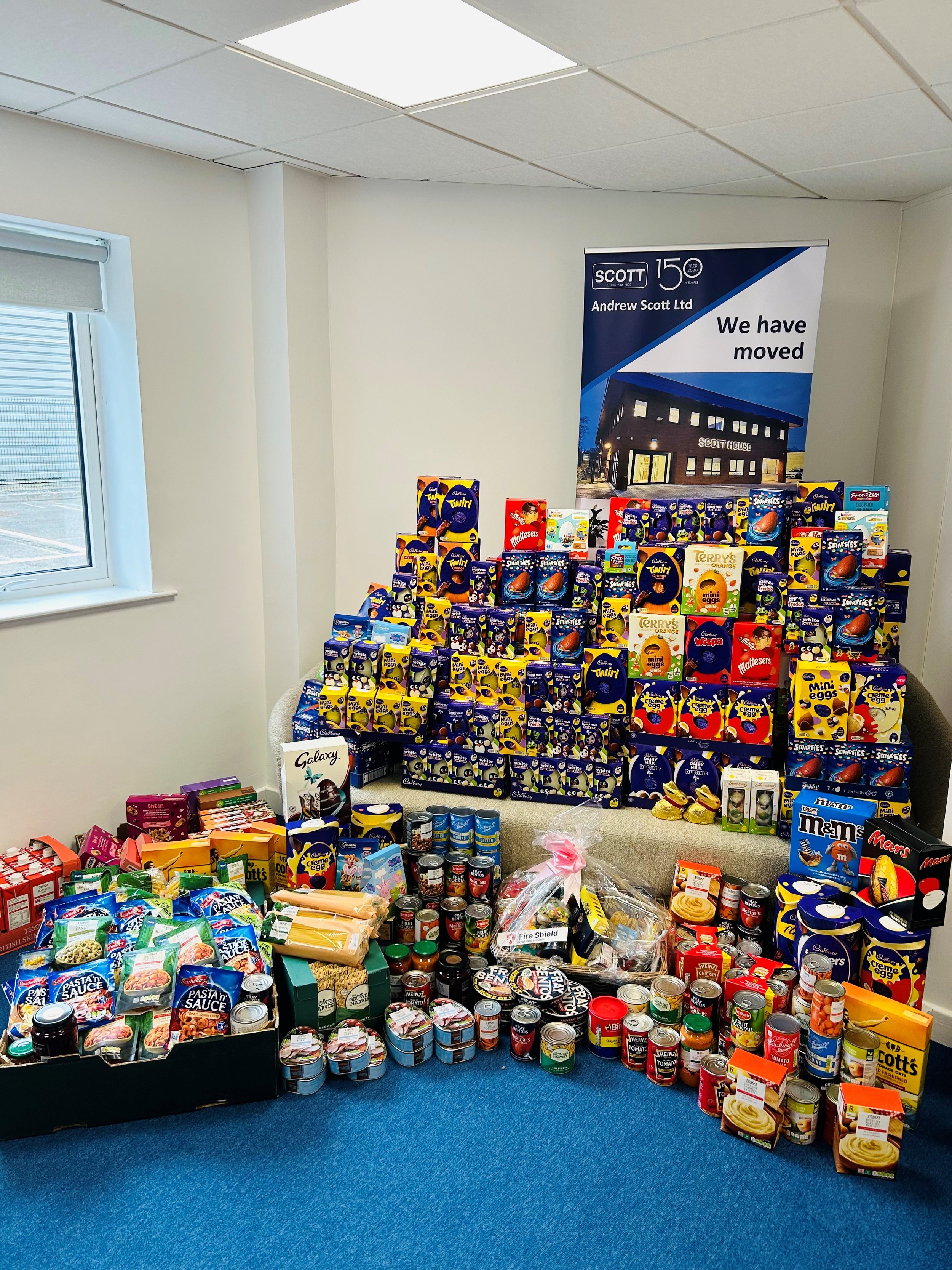 A room with a large display of assorted food and snack items, including boxes of chocolates, canned goods, pasta, and sauces, arranged on a bench and the floor. A sign in the background indicates the company has moved.