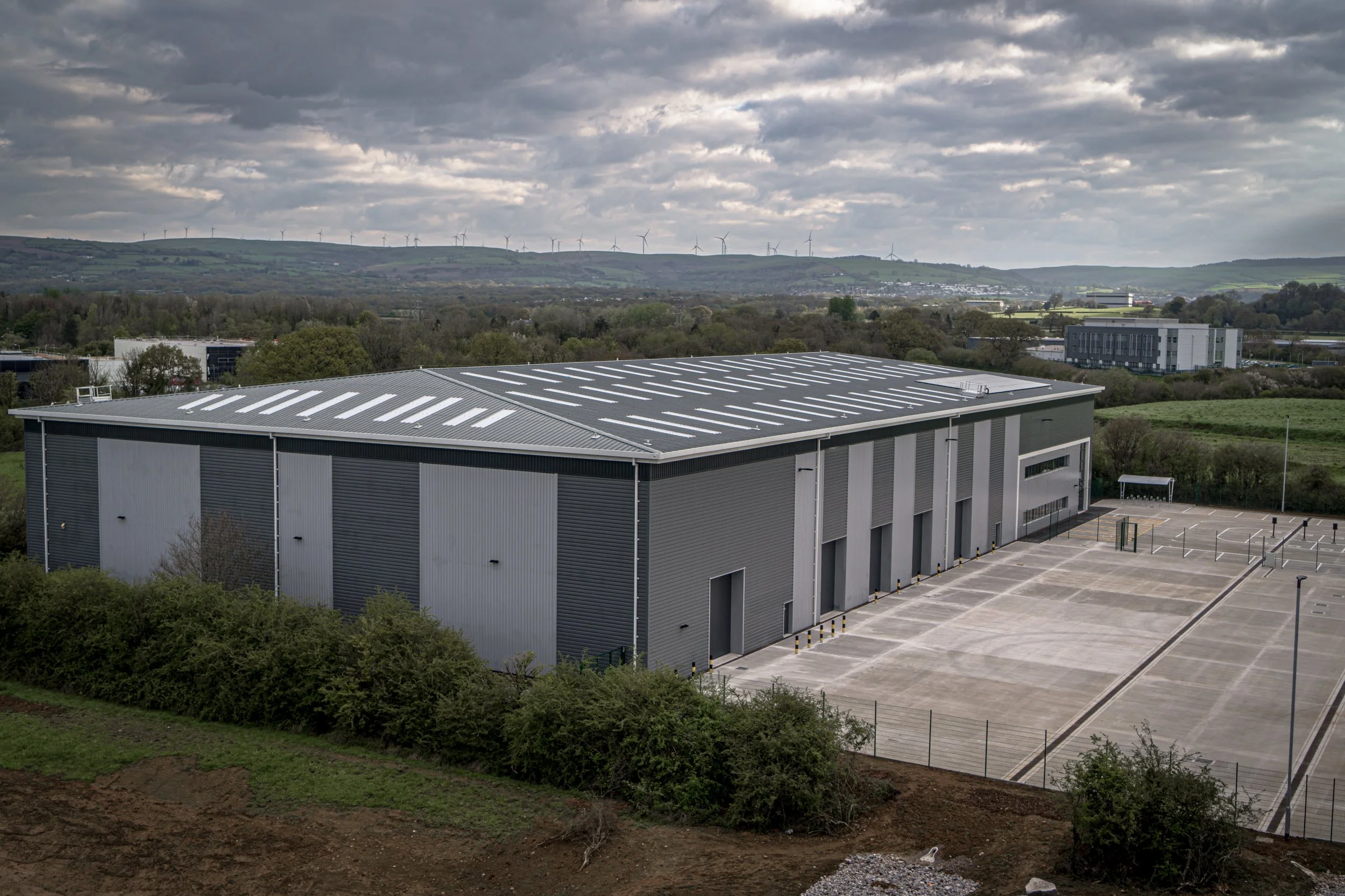 Large industrial warehouse with a parking lot, surrounded by greenery and hills in the background under cloudy skies.