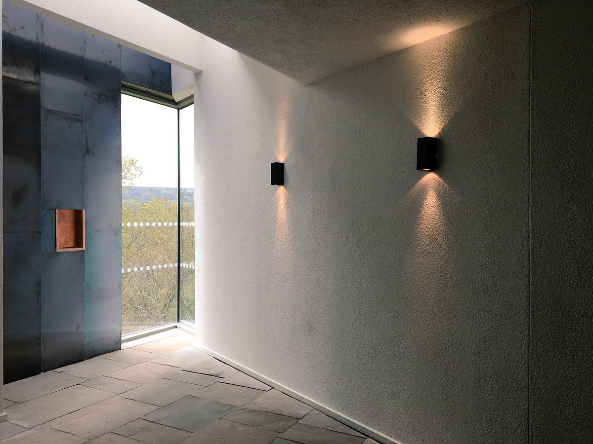 Interior corner with white textured wall, two black wall sconces casting upward and downward light, a large window revealing trees outside, and a section of dark metal paneling with a small copper-colored niche.