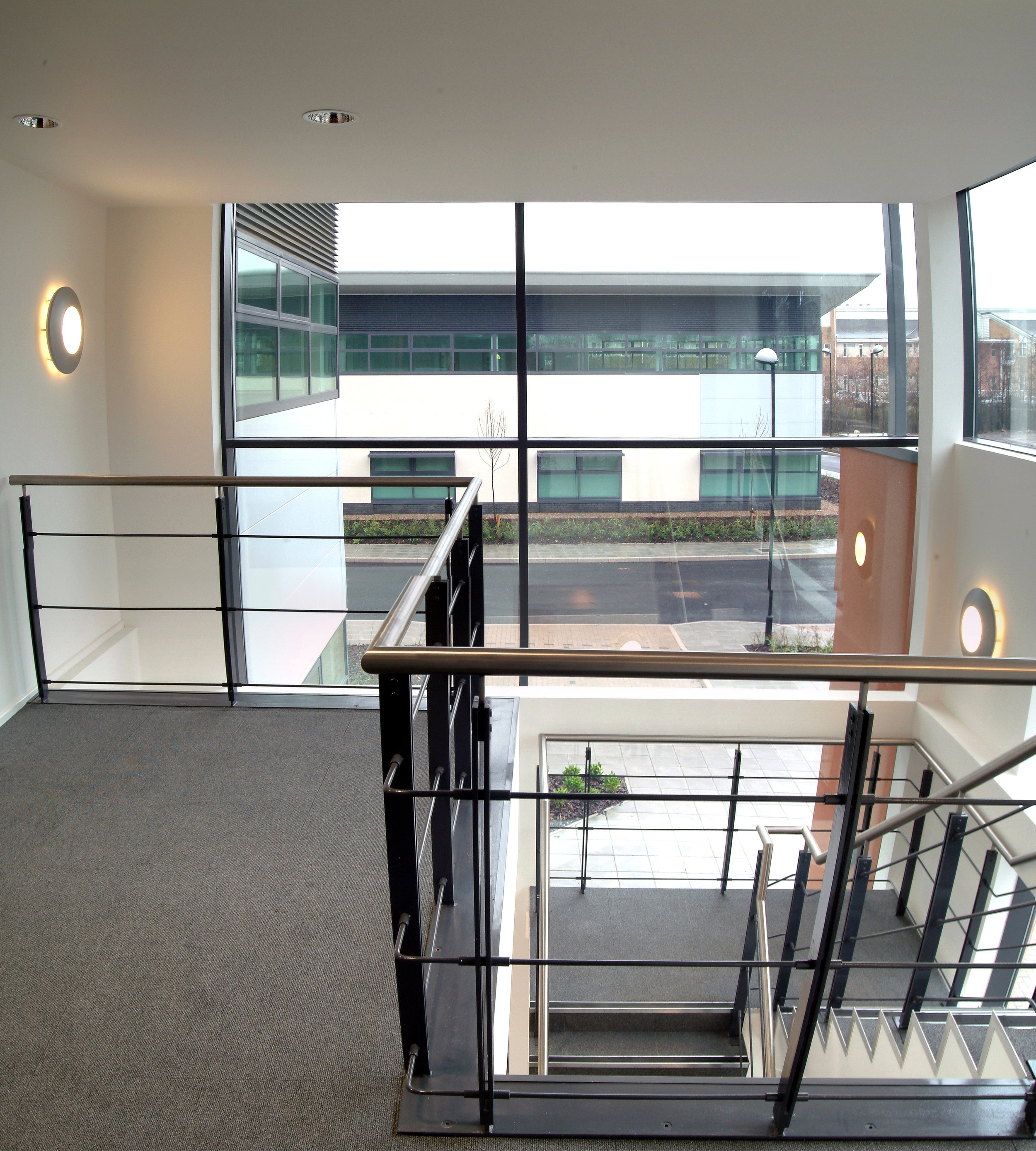 Interior view of a modern stairwell with metal handrails, gray carpeted flooring, and large panoramic windows overlooking the street outside.