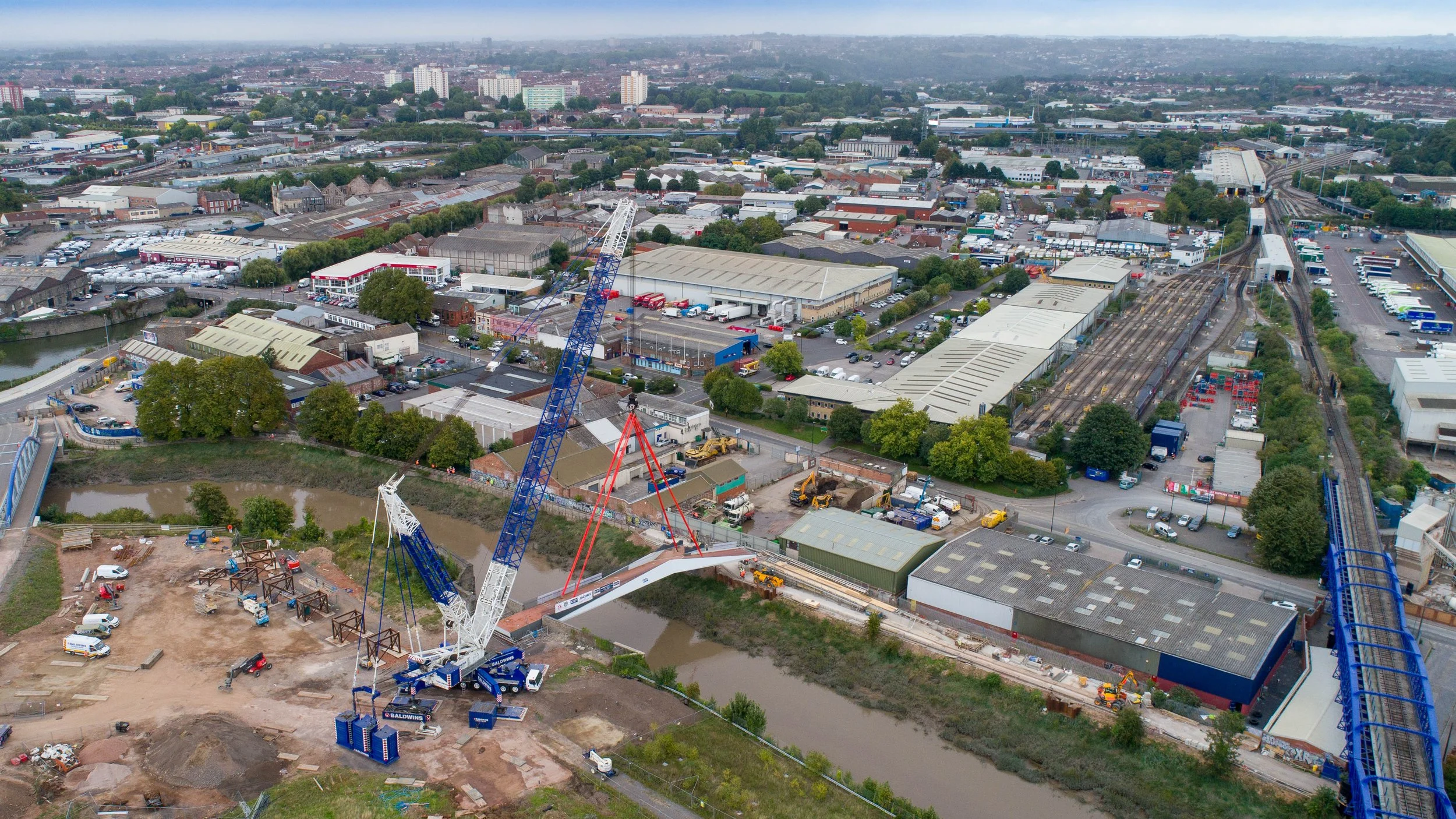 An aerial view of a construction site with cranes building a bridge over a small river in an urban area.