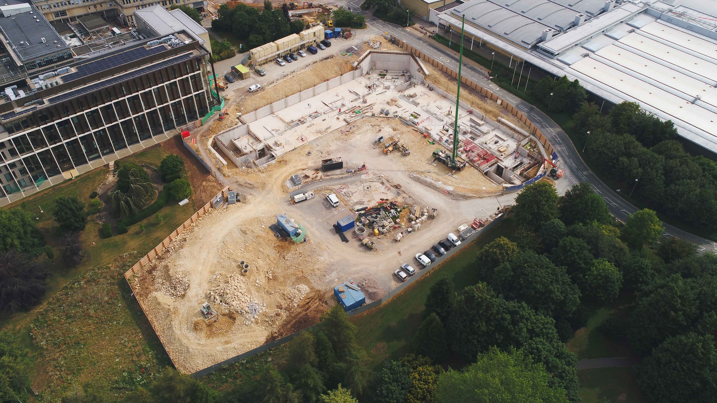 Aerial view of a construction site with cranes, building foundations, and scattered construction materials, situated next to a modern glass building and surrounded by trees and parked cars.