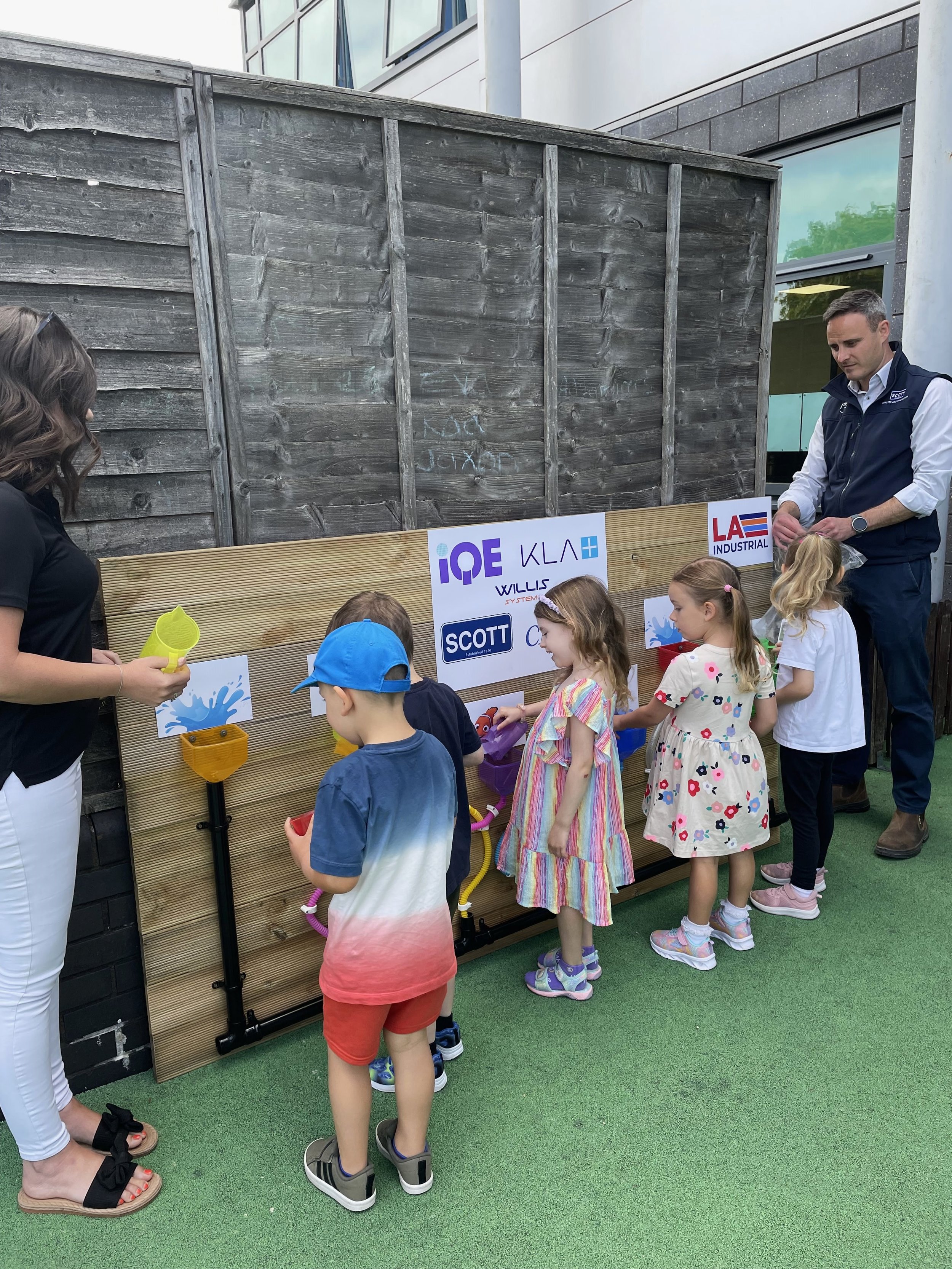 Children playing with water toys at a sensory activity station outside, assisted by adults, with a wooden backdrop and building in the background.