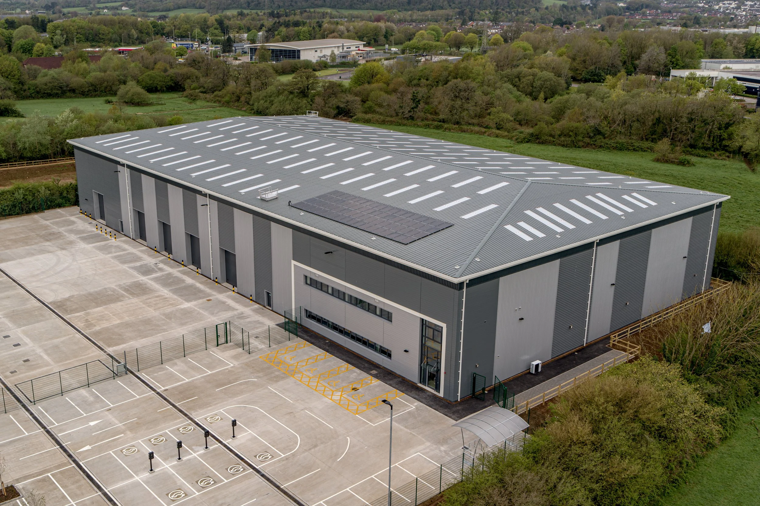 A large industrial building with a metal roof, solar panels, and a parking lot with multiple handicapped parking spaces, surrounded by greenery and trees.