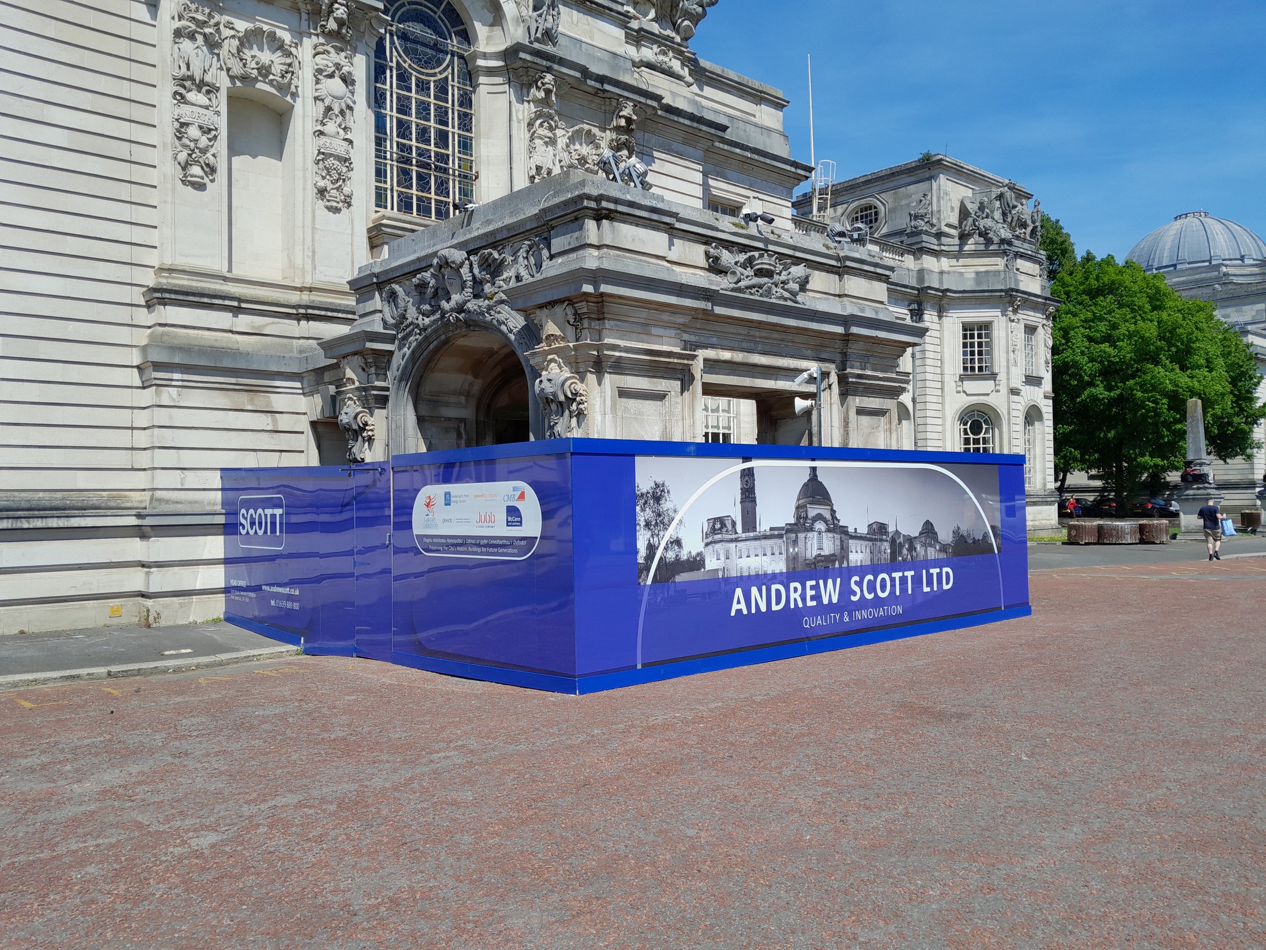 Construction barriers in front of a historic building with ornate stonework and large windows, featuring a blue sign with the name "Andrew Scott Ltd" and its logo, along with a black and white photograph of the building on the barrier.