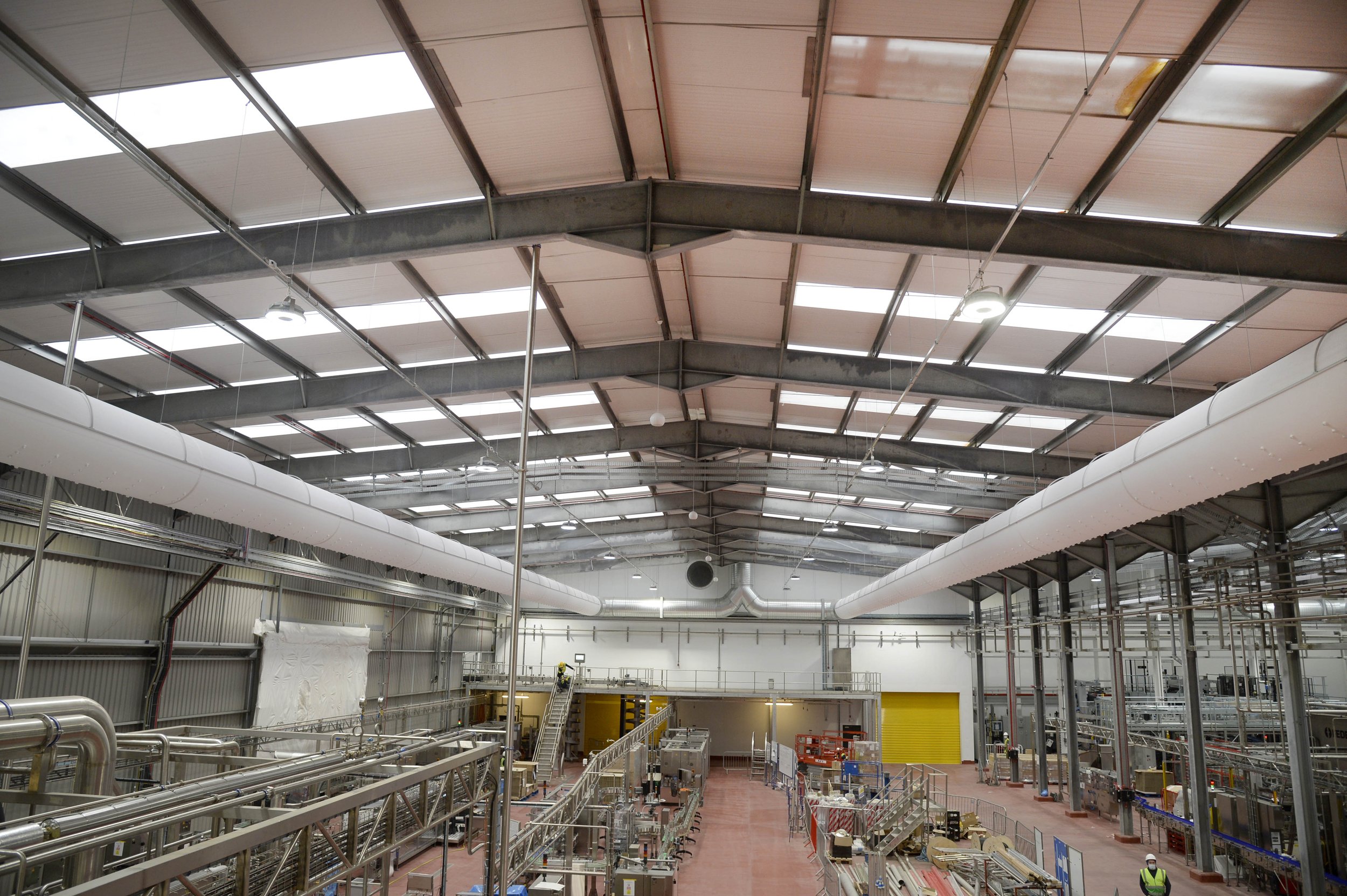 Interior view of an industrial warehouse or manufacturing facility with high ceilings, metal beams, ventilation ducts, and machinery.