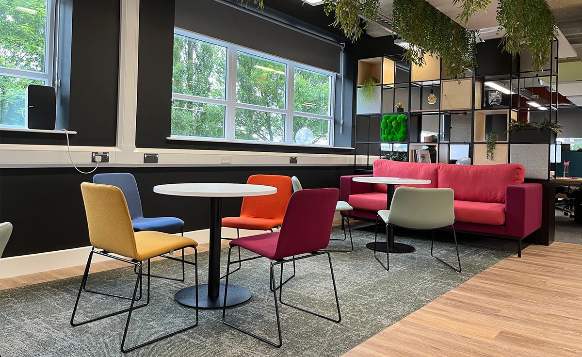 Modern office lounge area with colorful chairs, tables, and a pink sofa next to large windows and a black shelving unit. Green plants are hanging from the ceiling.