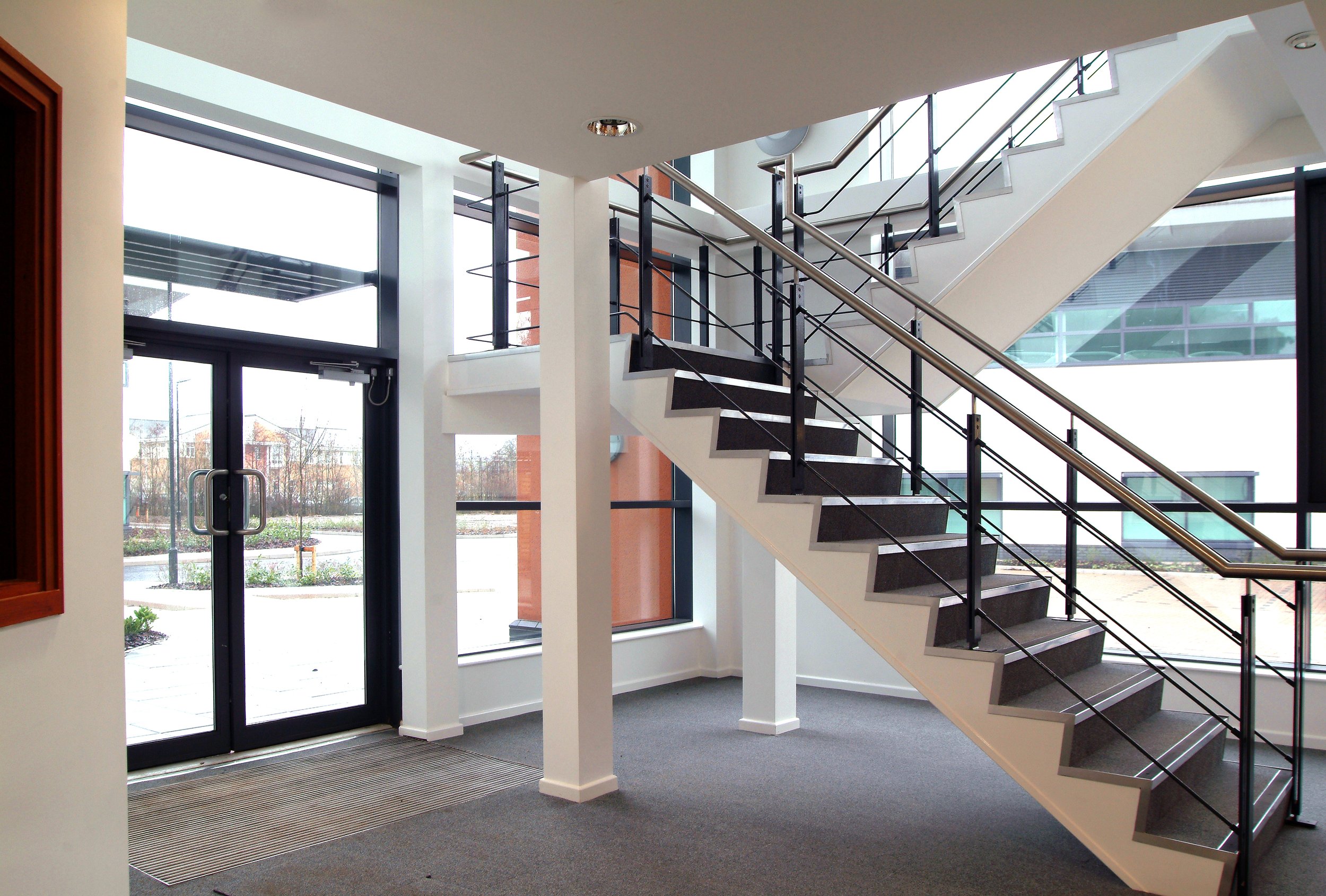 Interior of a modern building lobby with a staircase featuring black railings and dark steps, large glass windows, and a glass door entrance, with a view of outdoor landscaping.