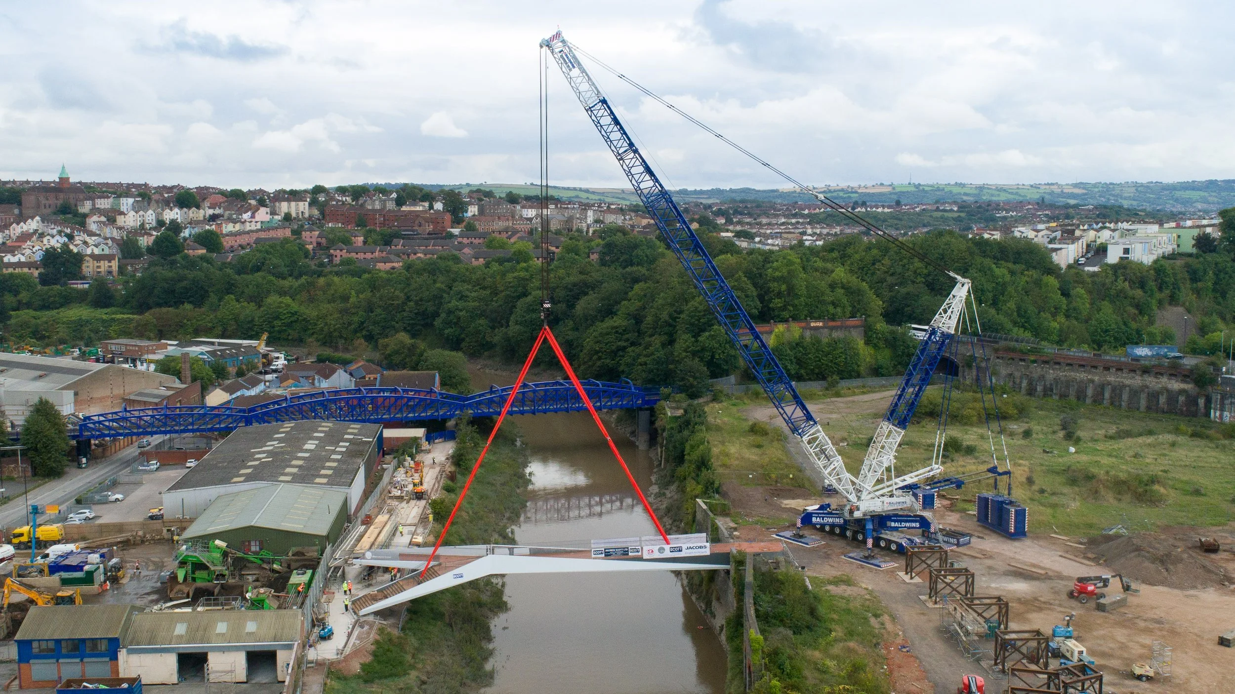 A large crane is installing a new bridge over a river, with a blue arch and white supports, in a construction site on the riverbank surrounded by industrial buildings and residential areas in the background.