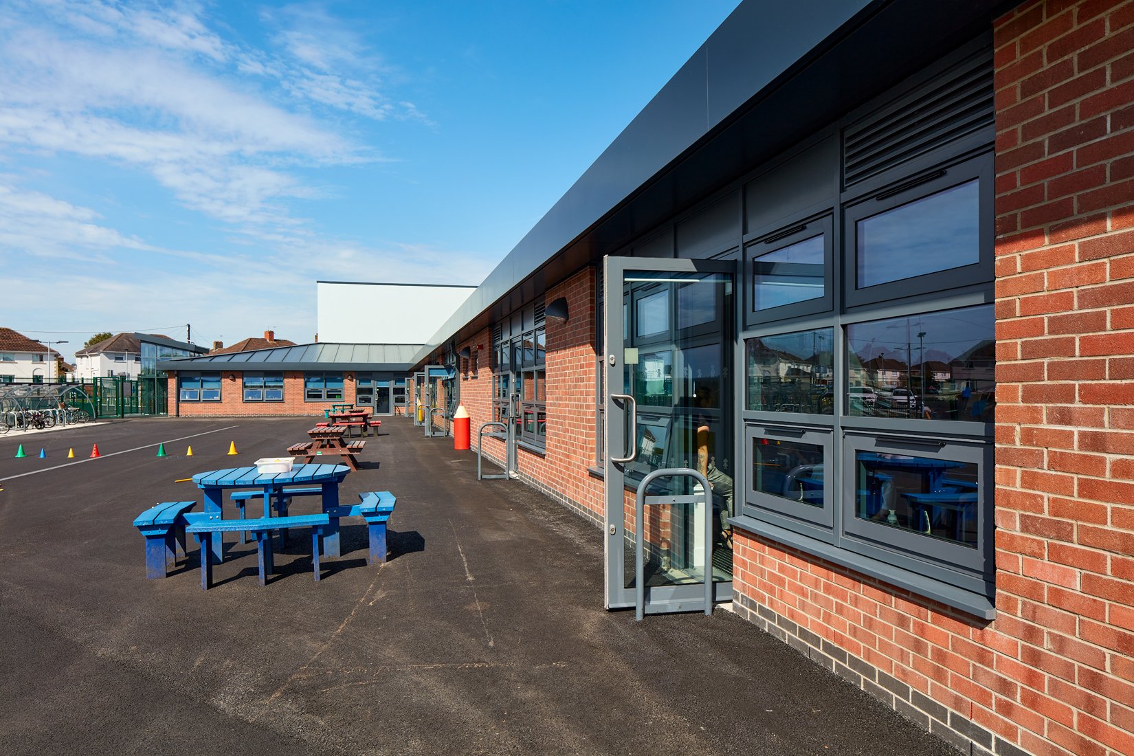 Open door leading to a schoolyard with colorful picnic tables and traffic cones on a sunny day.