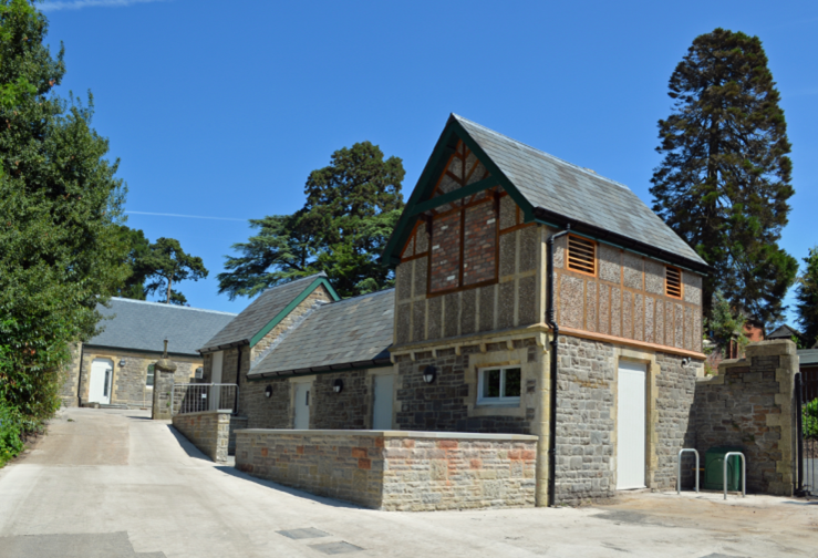 Residential house with stone and timber construction, pitched roof, and a small fence, surrounded by trees under a clear blue sky.