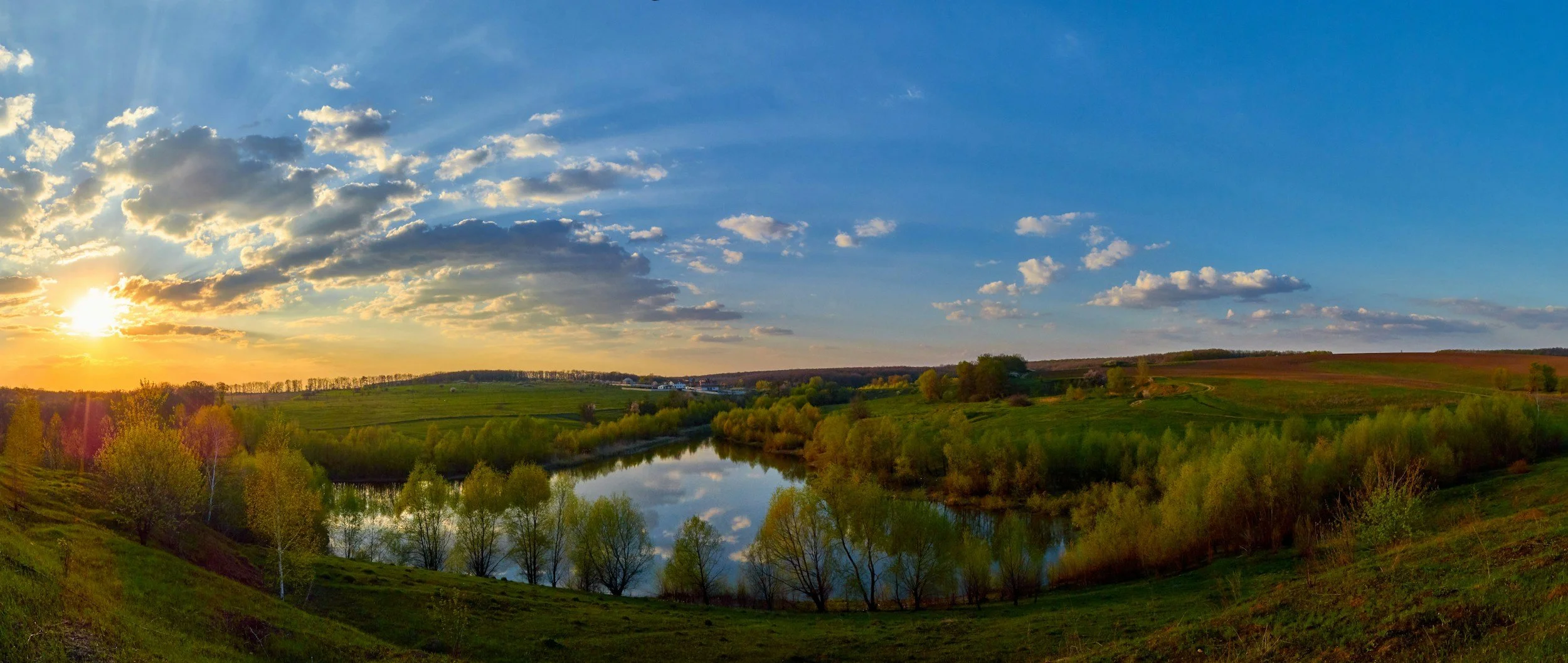 Paisaje con río, árboles verdes y colinas, sol en el horizonte con nubes en el cielo azul.